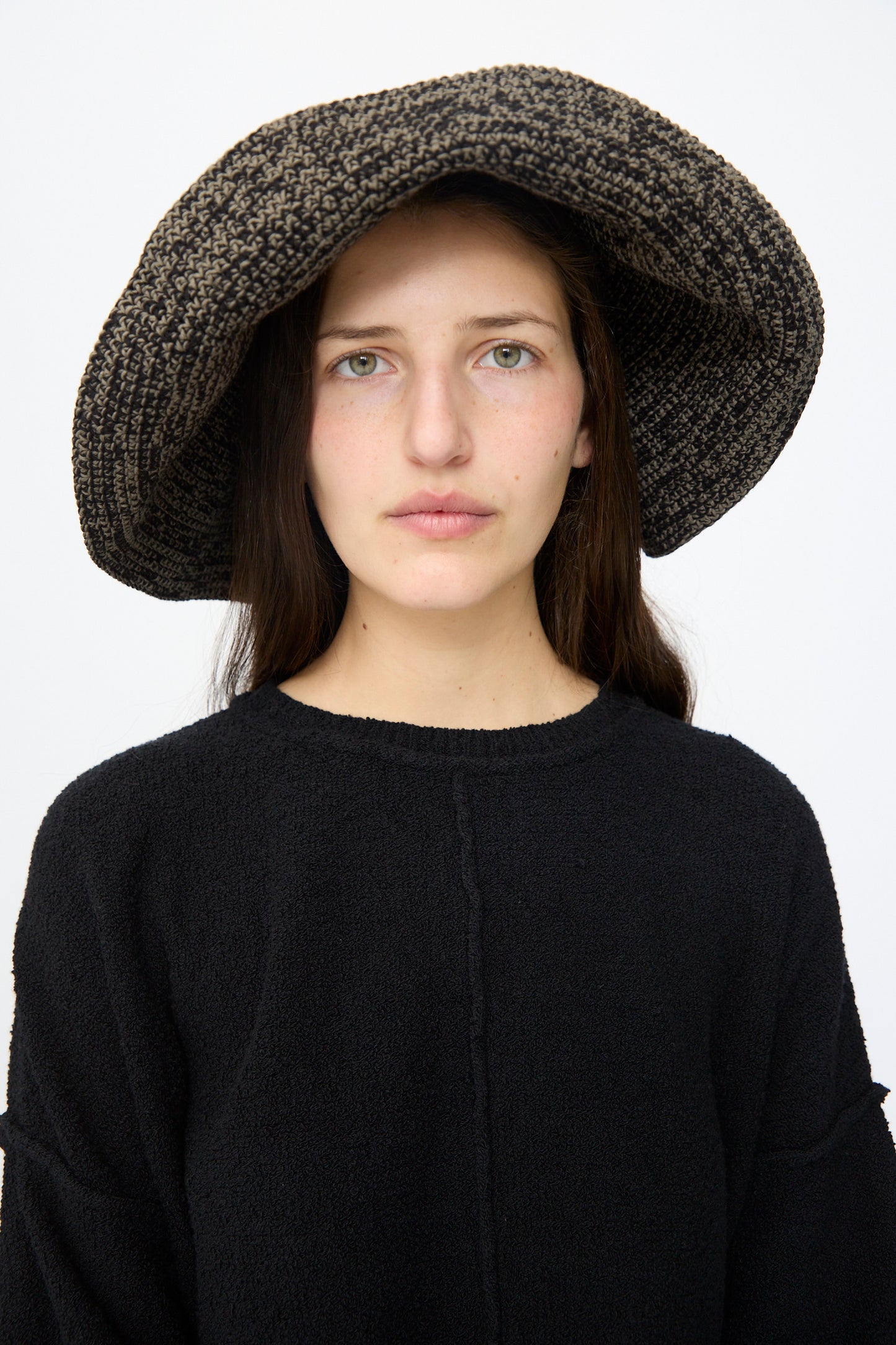 A woman with long brown hair wears the Lauren Manoogian Crochet Big Brim Hat in Black Marl and a matching sweater, standing against a plain white background.