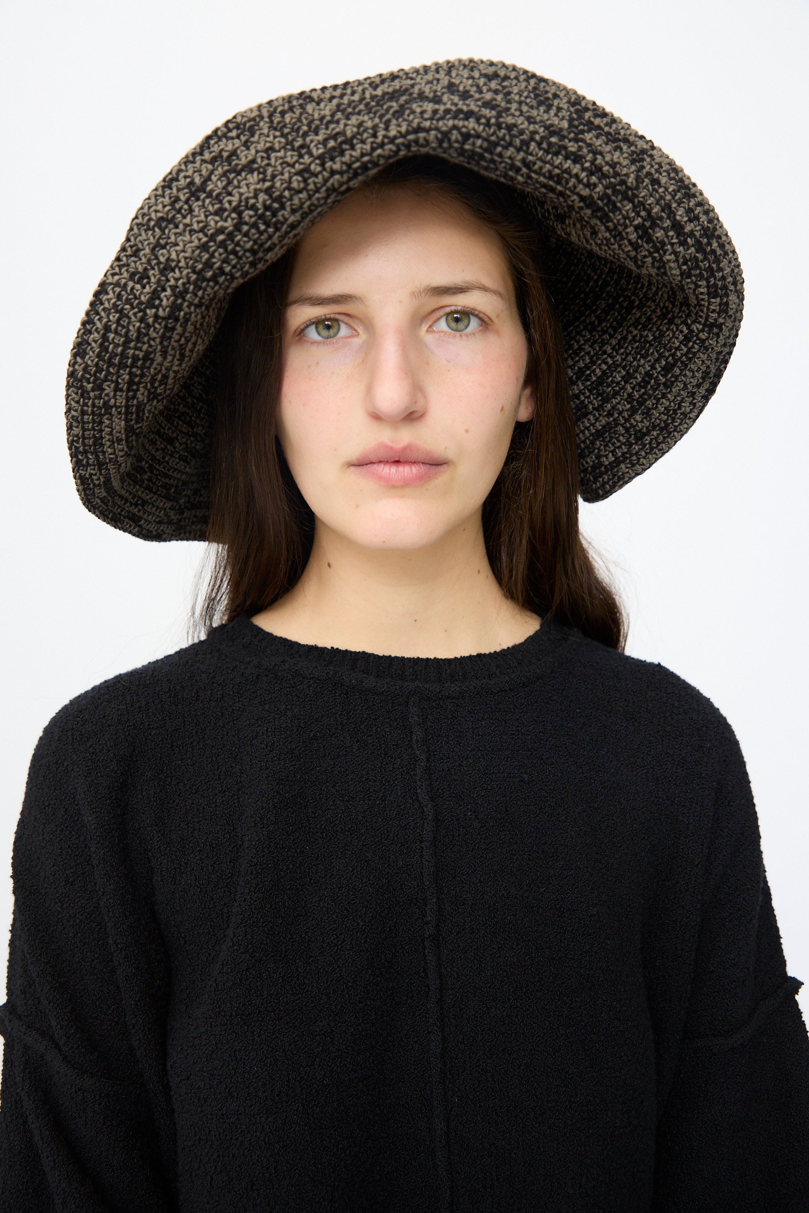 A woman with long brown hair wears the Lauren Manoogian Crochet Big Brim Hat in Black Marl and a matching sweater, standing against a plain white background.