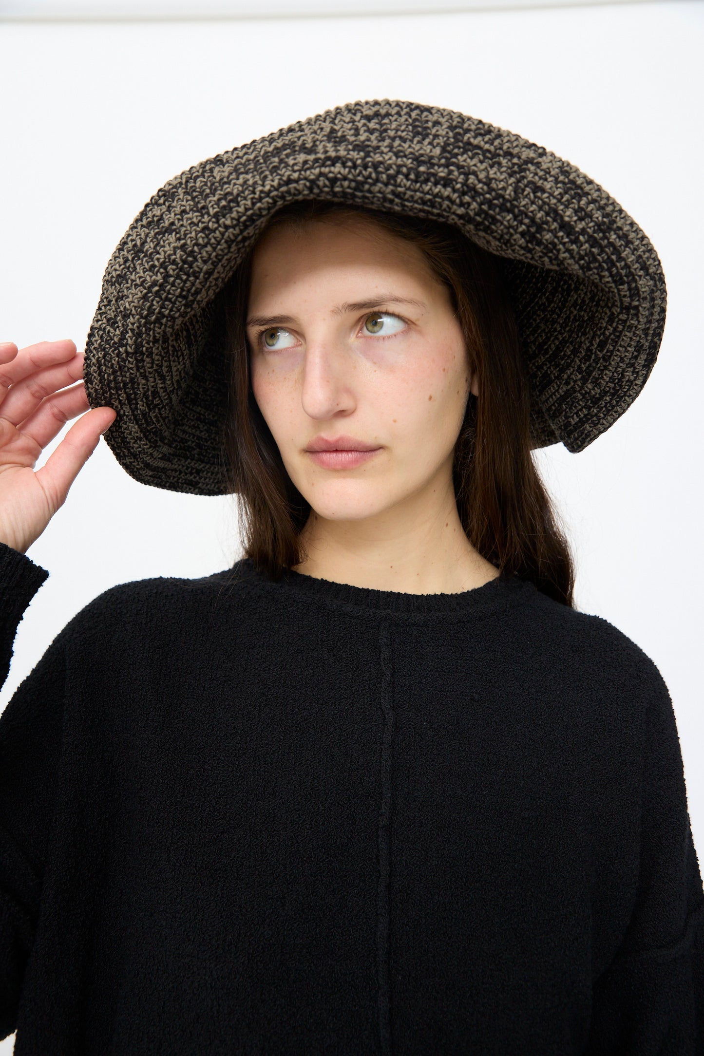 A woman in a Lauren Manoogian Crochet Big Brim Hat in Black Marl and a matching sweater gazes sideways against a plain white background.