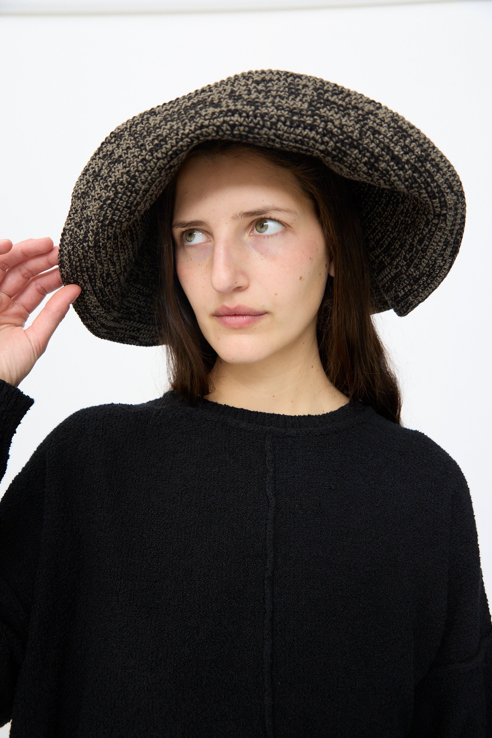 A woman in a Lauren Manoogian Crochet Big Brim Hat in Black Marl and a matching sweater gazes sideways against a plain white background.