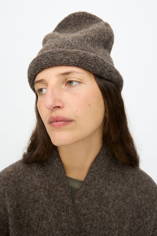 A person with long brown hair wears the Lauren Manoogian Crown Beanie in Patina and a matching sweater, looking slightly to the side with a neutral expression against a plain light background.