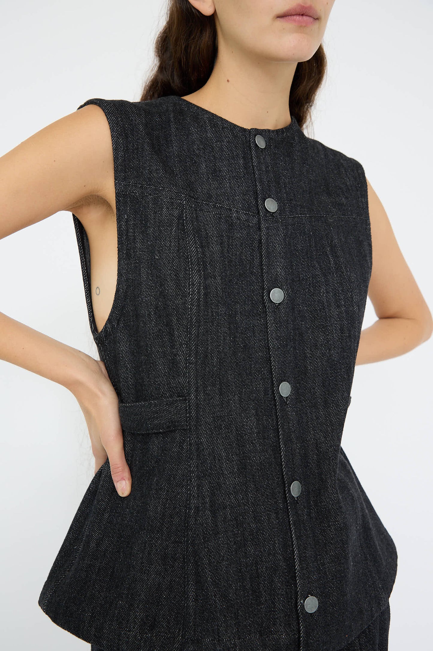 The woman confidently places her hands on her hips, wearing the sleeveless Denim Bodice in Indigo by Lauren Manoogian, a button-up top crafted from 100% cotton.