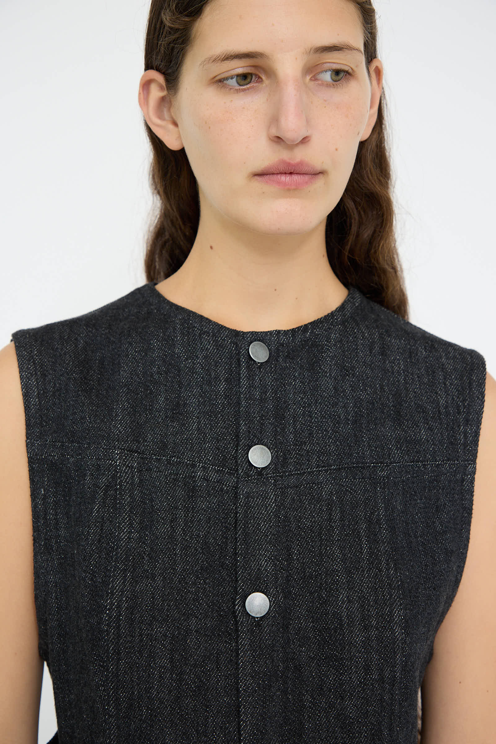 A person with long hair is wearing the Denim Bodice in Indigo, a sleeveless dark gray button-up top made from 100% cotton by Lauren Manoogian, looking to the side against a plain background.