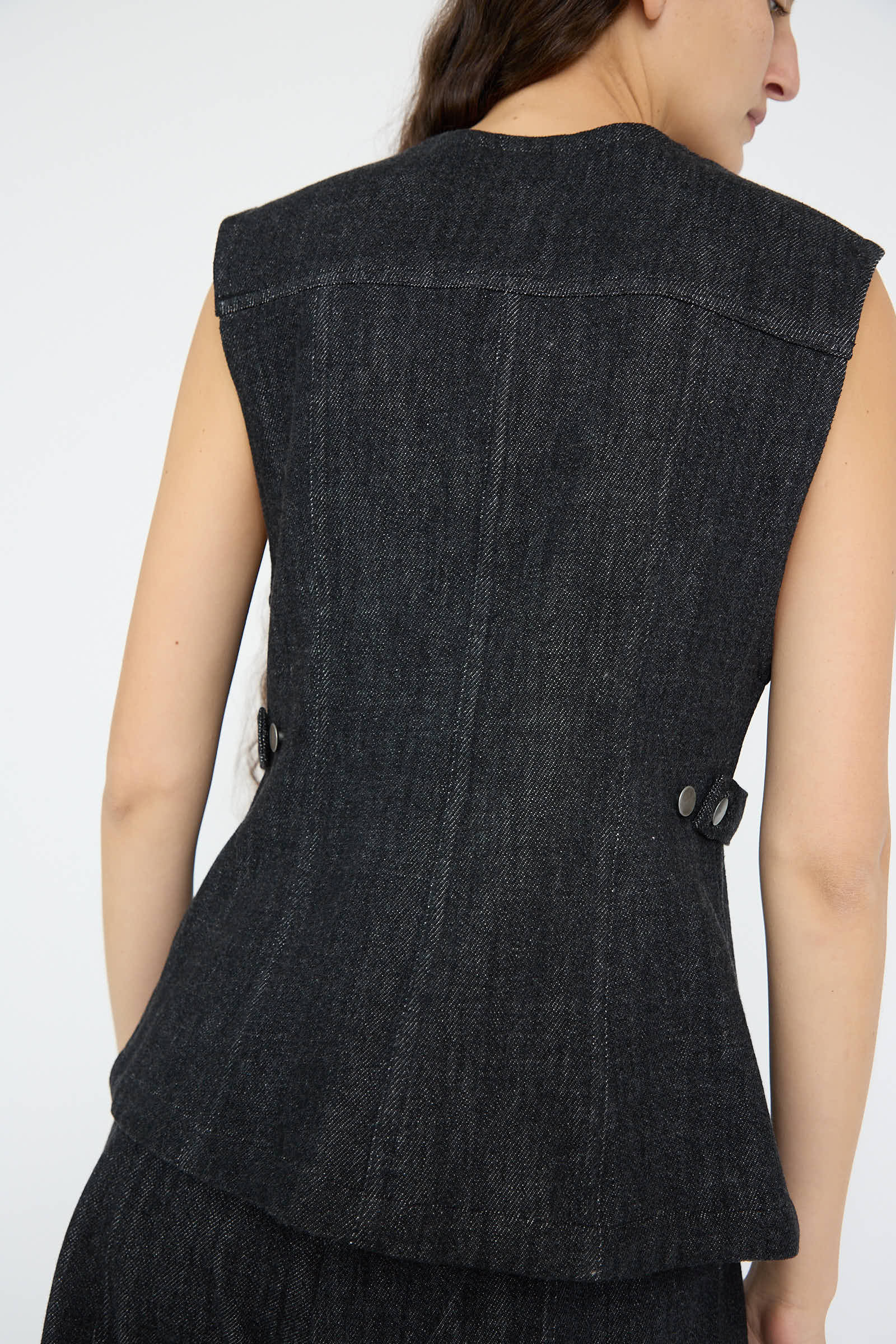 A person wearing the Lauren Manoogian Denim Bodice in Indigo, a sleeveless jacket crafted from 100% cotton with button details on the back and an adjustable waist for an impeccable fit.