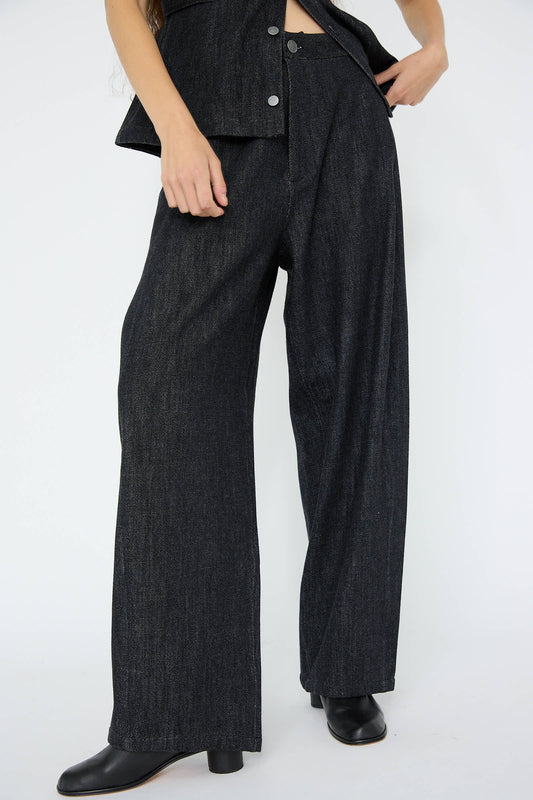 A person stands against a plain white background, wearing the Denim Pant in Indigo from Lauren Manoogian, featuring high-waisted, wide-leg trousers crafted from 100% cotton. The ensemble includes a matching dark top and black heeled boots, adding sophistication and comfort to the polished look.