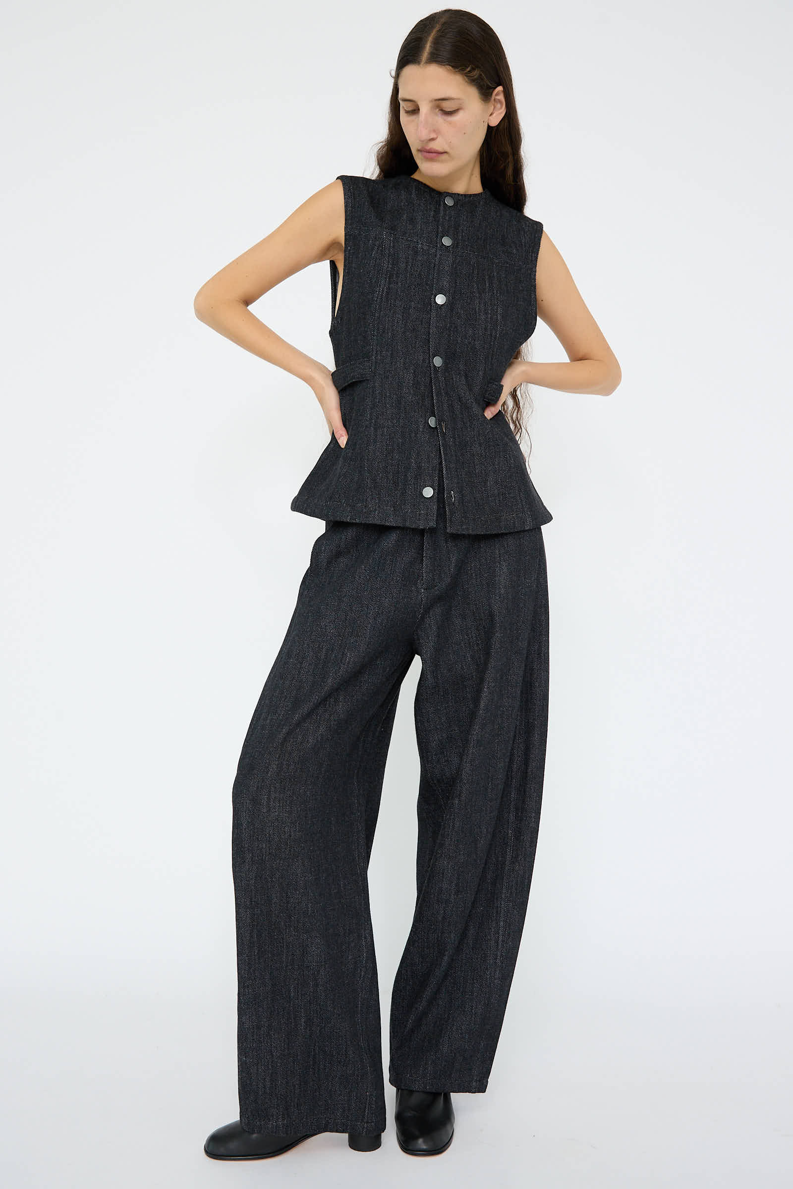 A person is posed against a plain background, wearing a sleeveless button-up top paired with Lauren Manoogian's Denim Pant in Indigo, featuring wide-leg design and an adjustable waist for comfort.