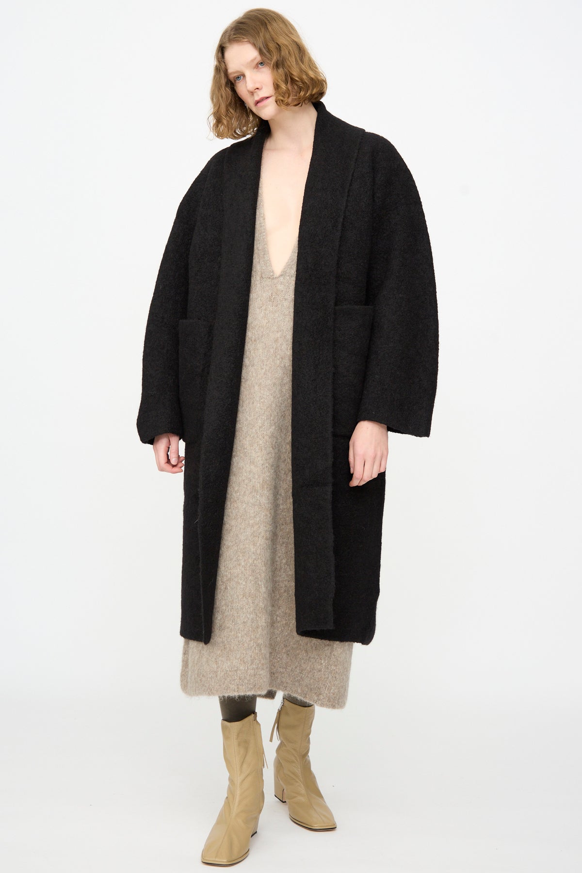 Wearing the Lauren Manoogian Double Face Long Coat in Black—crafted from alpaca wool in Peru—over a loose beige dress with tan boots, the person stands against a plain white background.