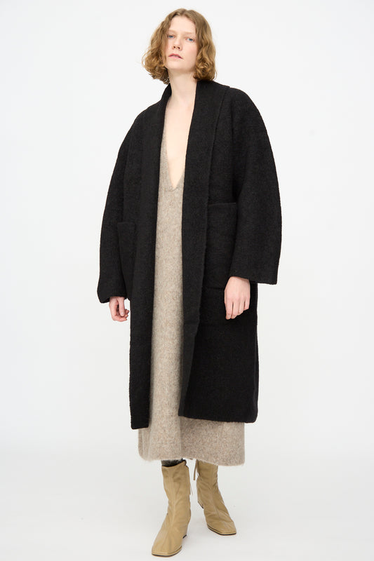 A person wearing the Lauren Manoogian Double Face Long Coat in Black, made from luxurious alpaca wool in Peru, layered over a beige dress and beige boots, stands against a plain white background.
