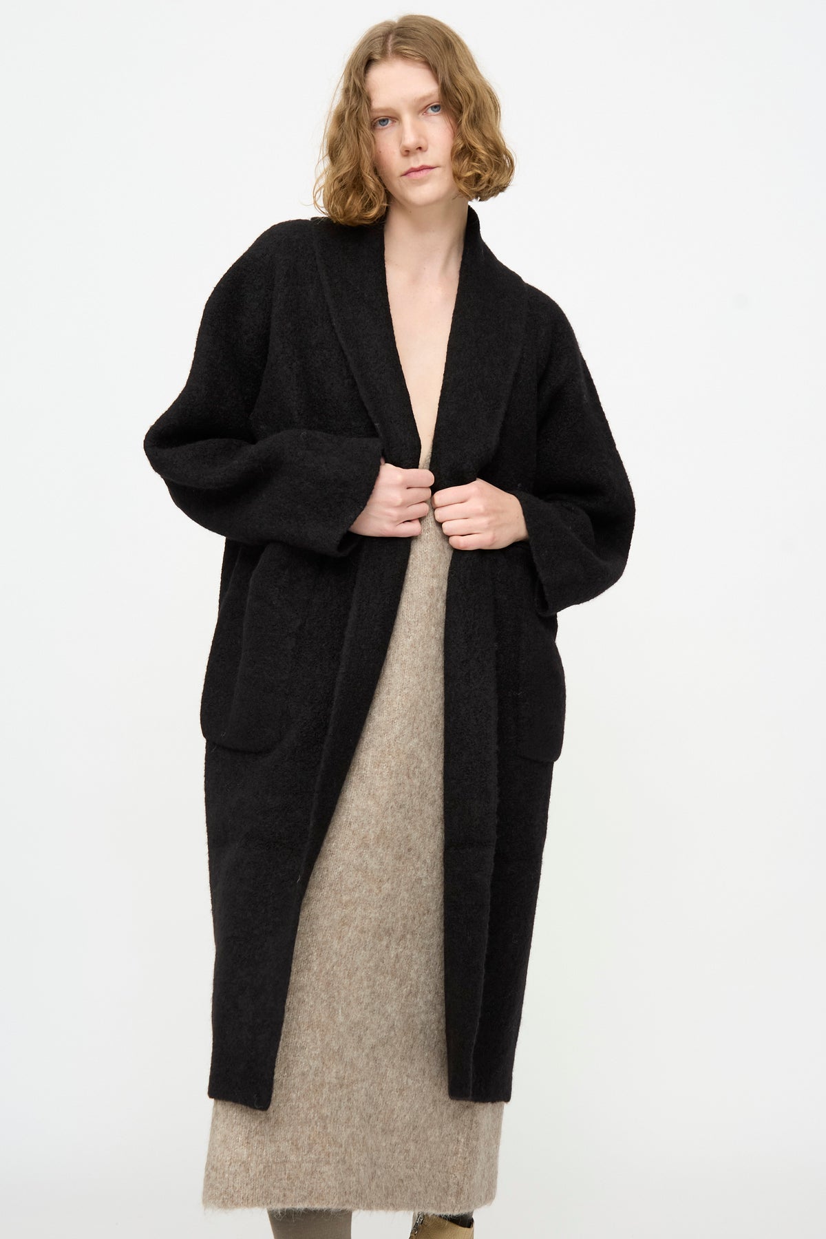 A person wearing the Lauren Manoogian Double Face Long Coat in Black, crafted from luxurious alpaca wool in Peru, layered over a beige textured dress. They stand against a plain white background, looking at the camera with hands holding the coat.