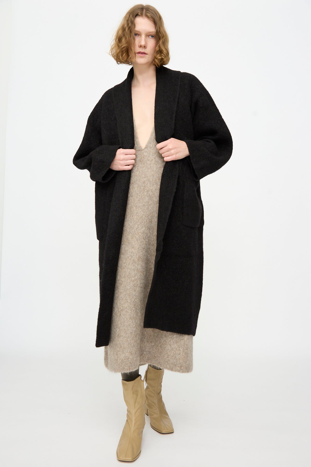 A person wearing the Lauren Manoogian Double Face Long Coat in Black, crafted from luxurious alpaca wool in Peru, stands against a plain white background, layered over a beige knit dress and beige boots.
