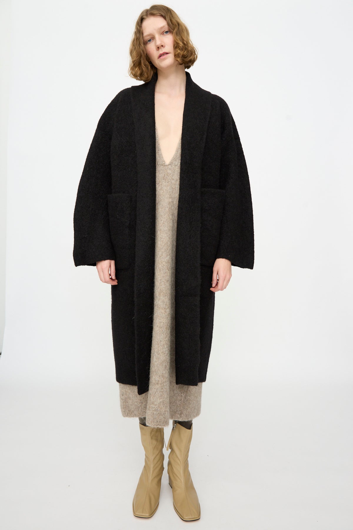 Person stands against a white background wearing the Lauren Manoogian Double Face Long Coat in Black—crafted from alpaca wool in Peru—over a beige dress and tan boots, with hands relaxed by their sides.