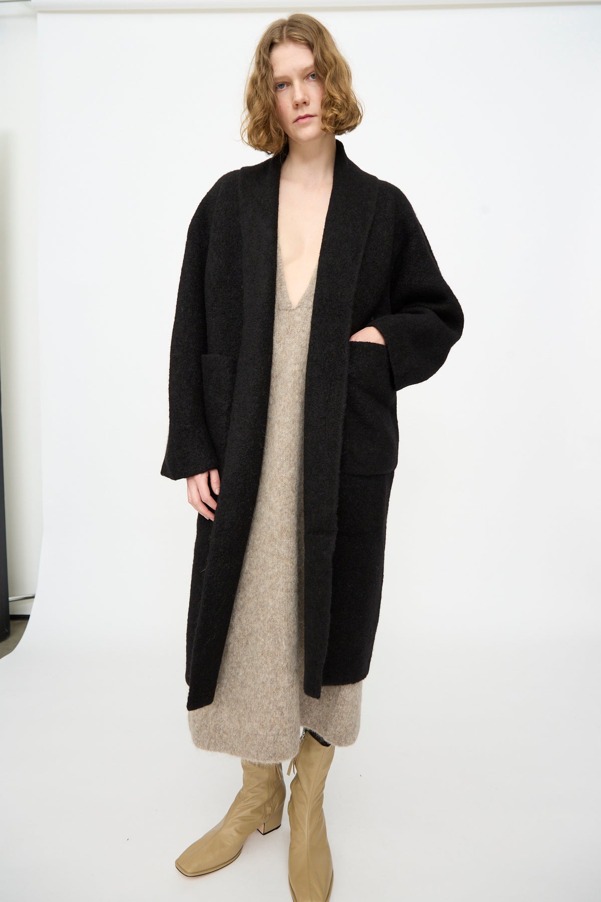 A person wearing the Lauren Manoogian Double Face Long Coat in Black—crafted in Peru from alpaca wool—layered over a beige knit dress and beige boots, standing against a plain white background.