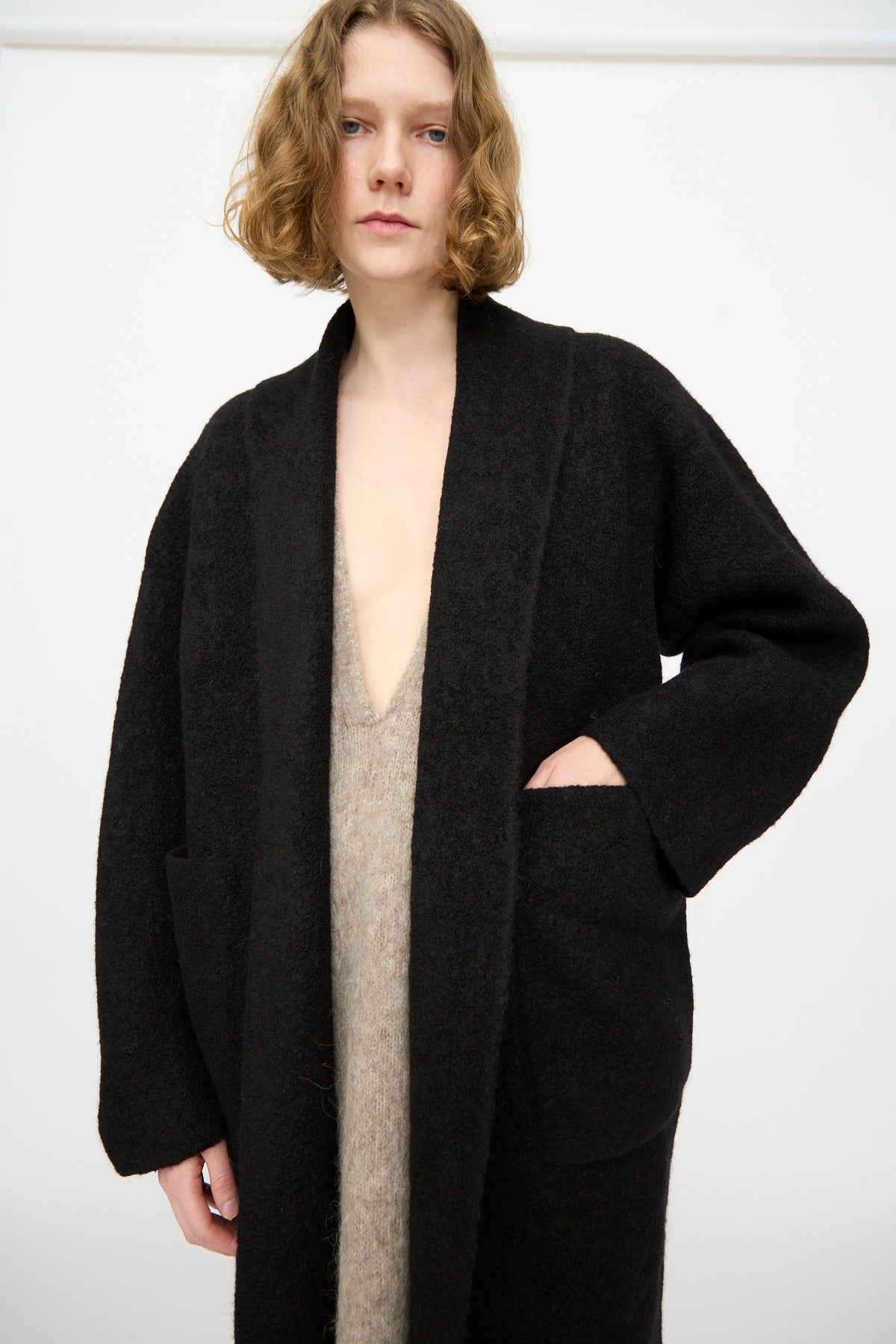 A person with shoulder-length blond hair wears the Lauren Manoogian Double Face Long Coat in Black, crafted from alpaca wool in Peru, layered over a beige V-neck garment and posed against a plain white background.