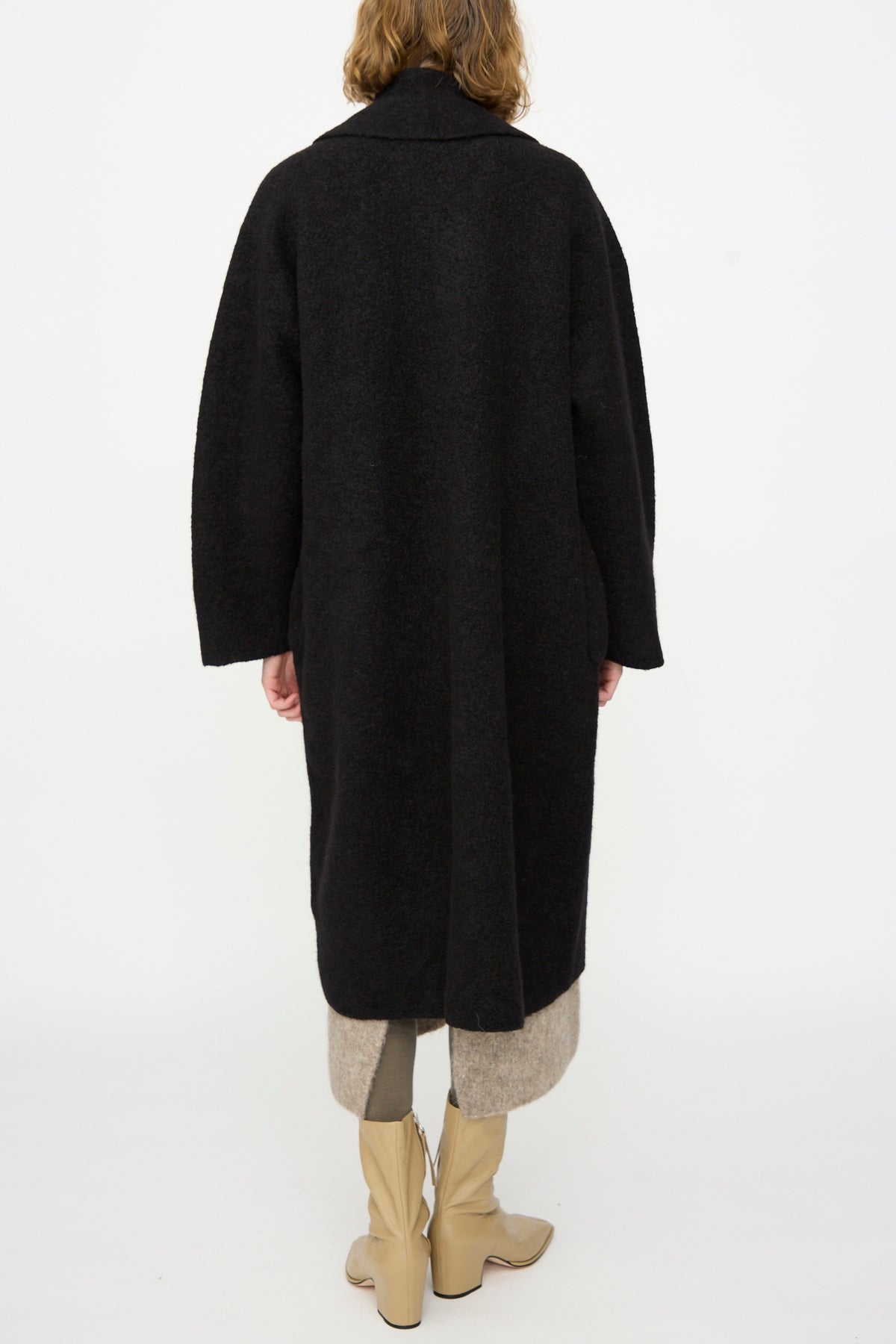 A person stands facing away on a plain white background, wearing the Lauren Manoogian Double Face Long Coat in Black—crafted from alpaca wool in Peru—layered over a beige skirt or pants, and paired with tan knee-high boots.