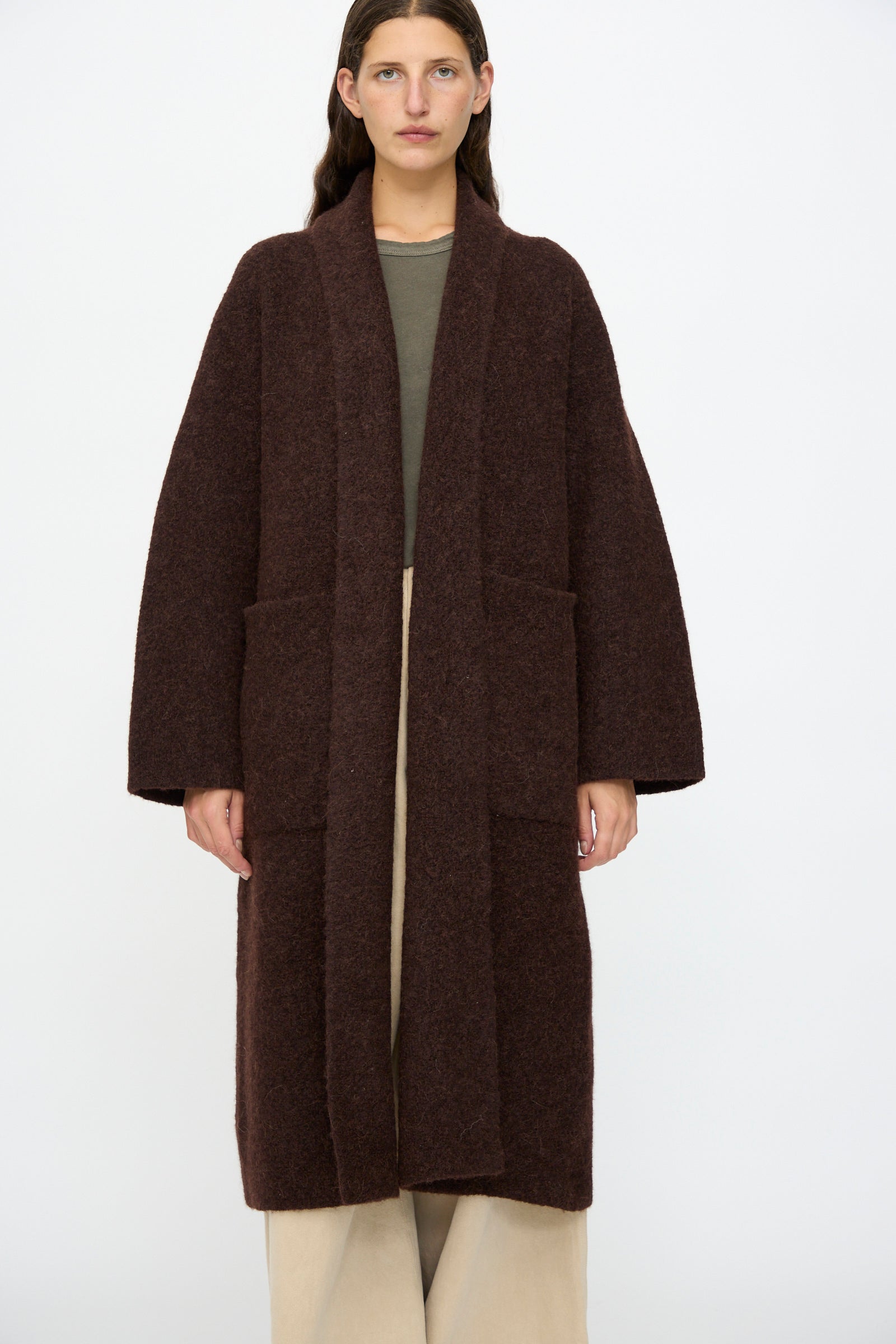 A person stands facing forward wearing the Lauren Manoogian Double Face Long Coat in Merlot over a green top and beige wide-leg pants, set against a plain white background.