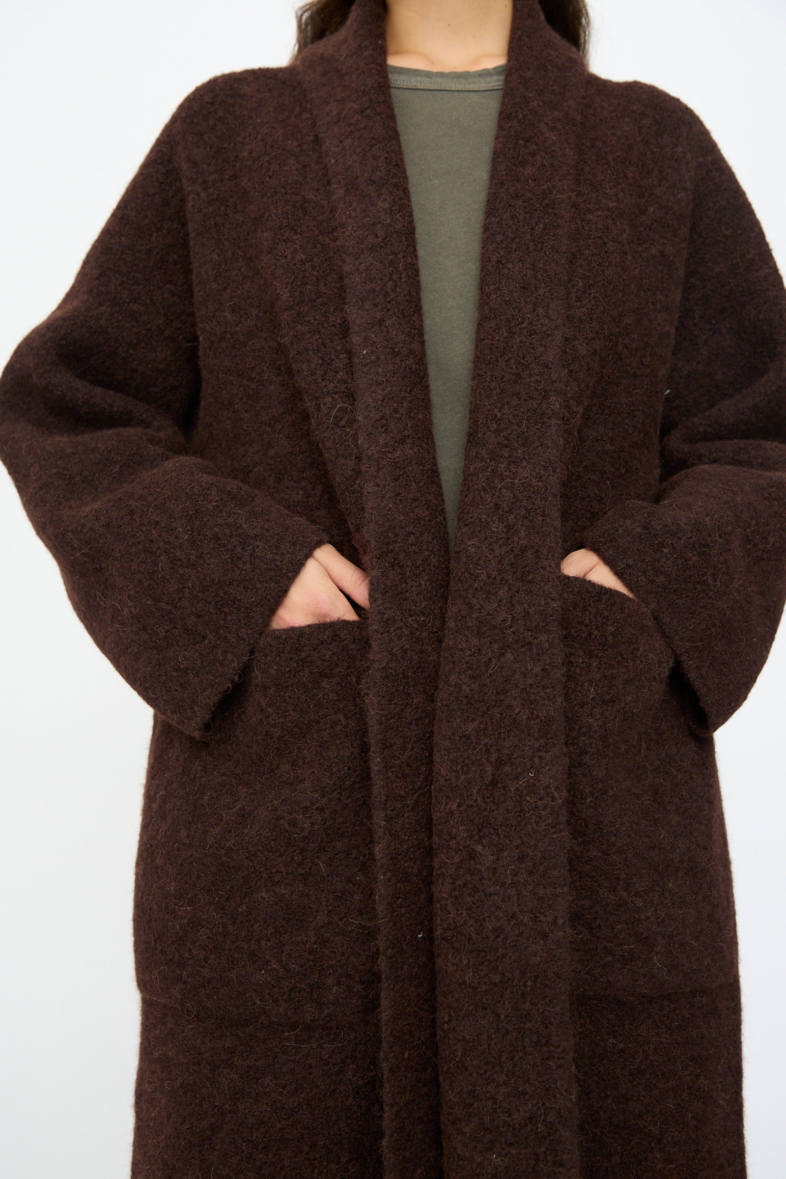 A person wearing Lauren Manoogian’s Double Face Long Coat in Merlot over a green top, with hands partially in the coat’s pockets.
