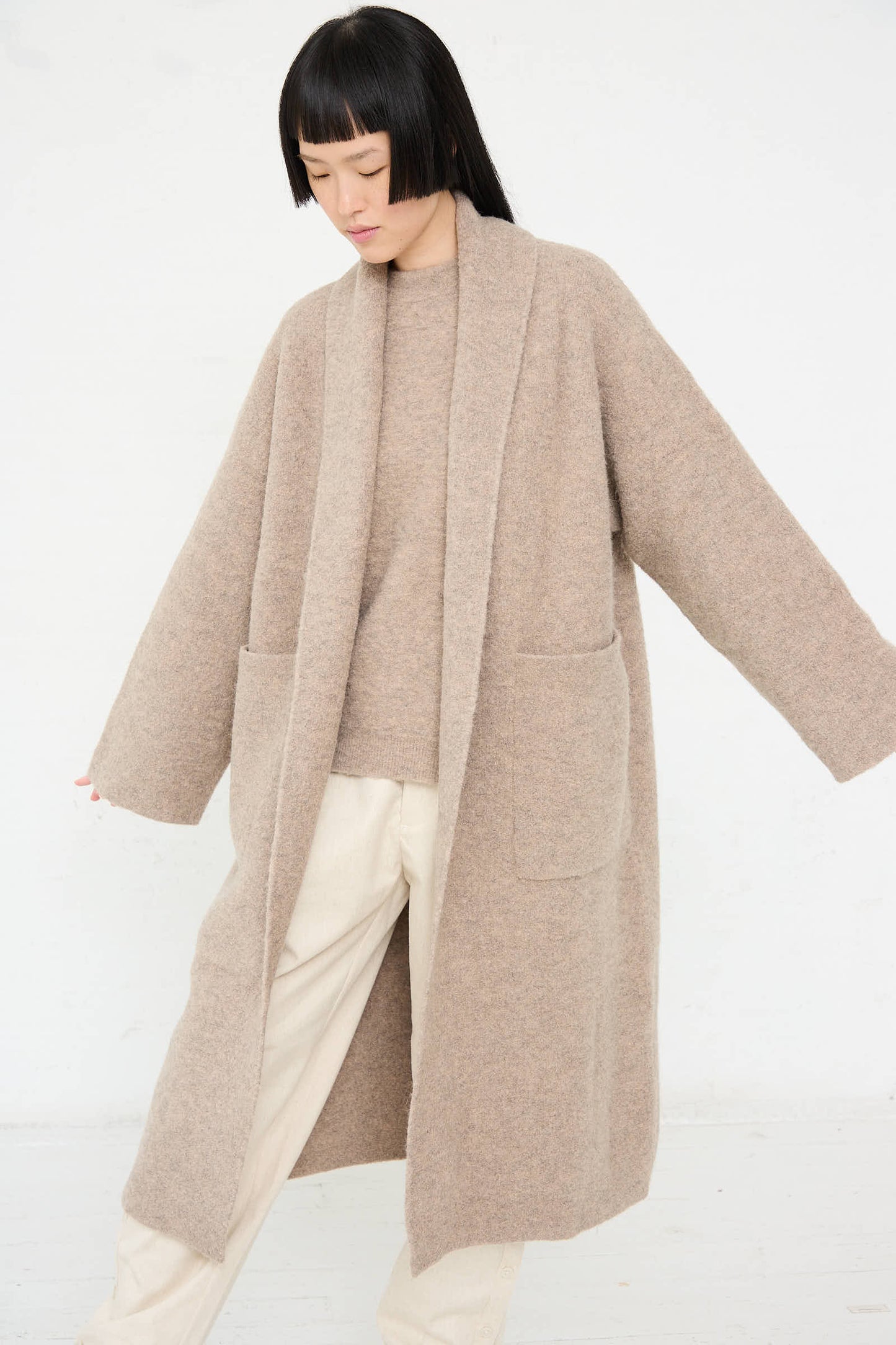 A person wearing an oversized Double Face Long Coat in Moth by Lauren Manoogian, featuring oversized patch pockets, and light-colored pants stands with arms slightly extended against a plain white background.