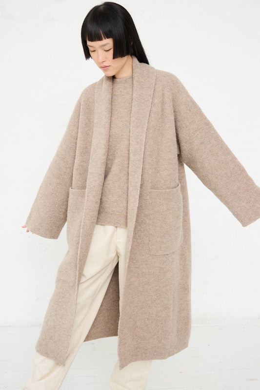 A person wearing an oversized Double Face Long Coat in Moth by Lauren Manoogian, featuring oversized patch pockets, and light-colored pants stands with arms slightly extended against a plain white background.