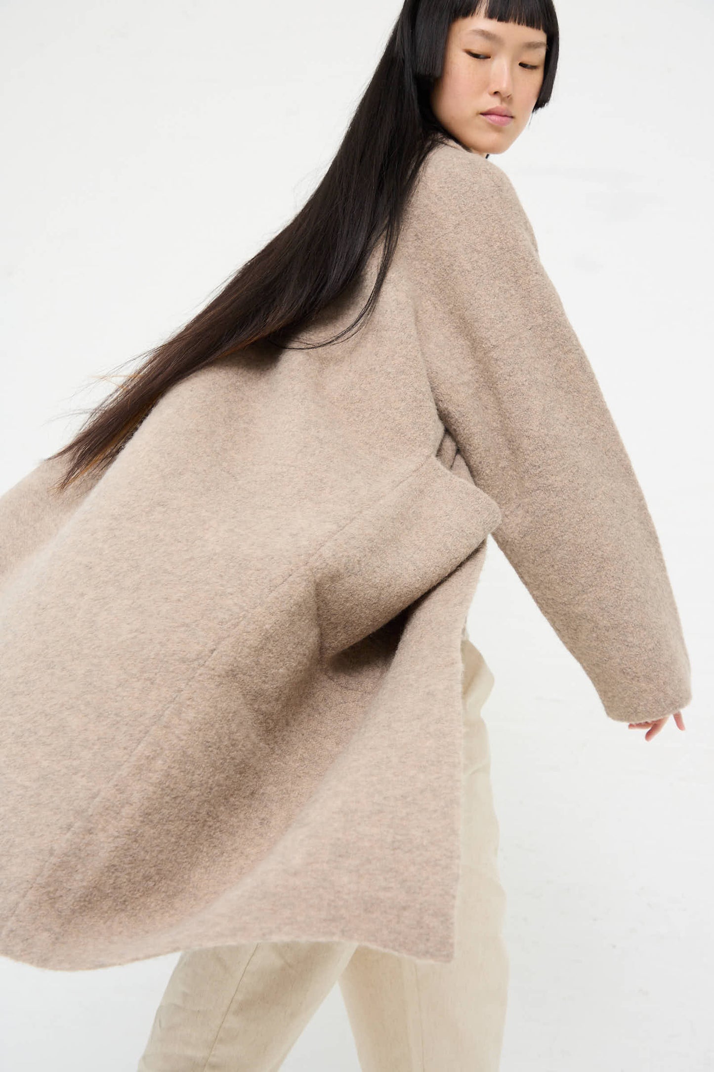 A person with long dark hair is wearing the Lauren Manoogian Double Face Long Coat in Moth, featuring oversized patch pockets and long sleeves, along with light-colored pants, facing sideways against a plain white background.