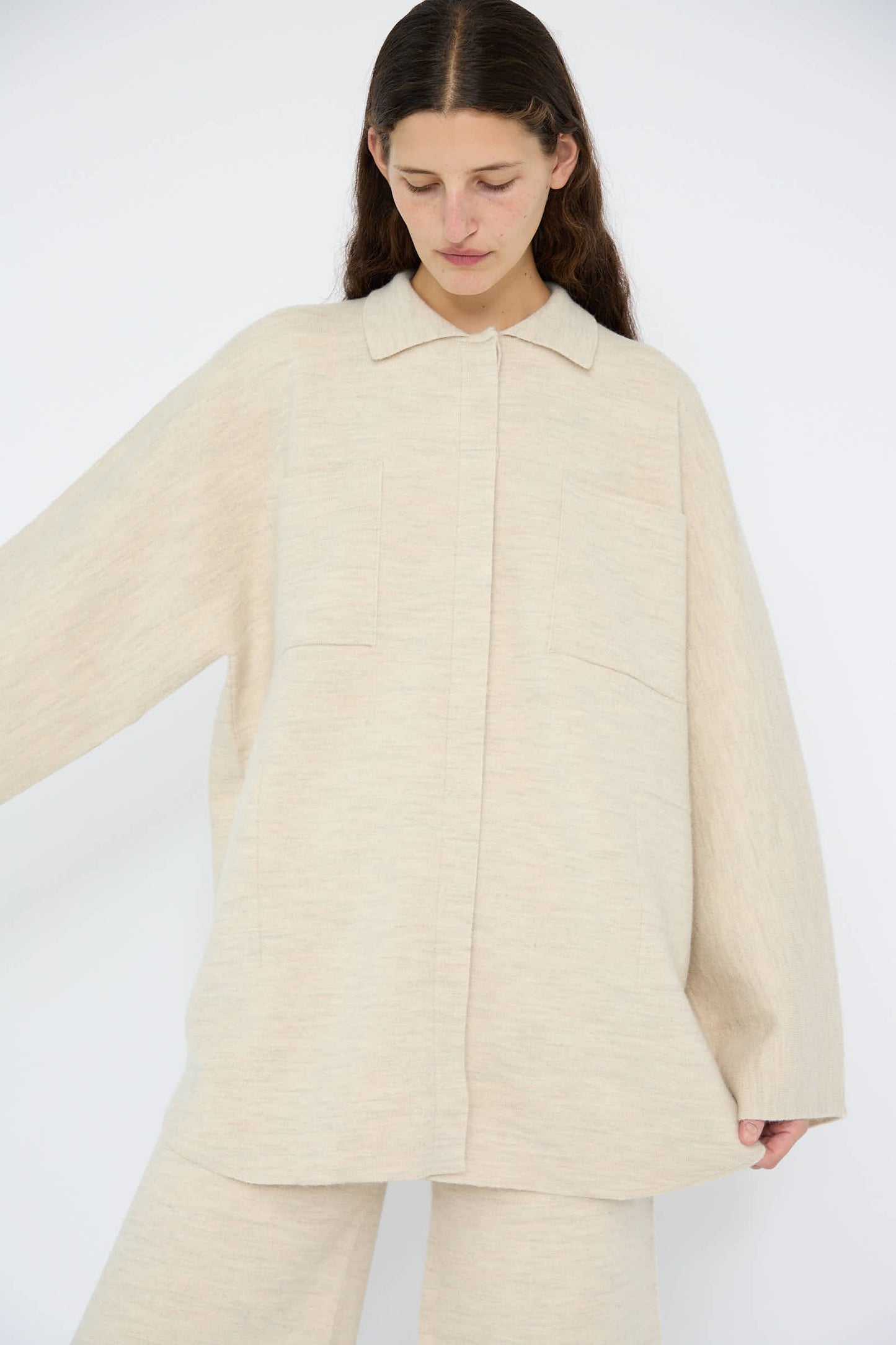 The individual is wearing the Lauren Manoogian Double Knit Big Shirt in Ecru, an oversized beige ensemble with wide sleeves and a cotton blend collared shirt, expertly crafted in Peru. They are gazing downwards against a plain background.