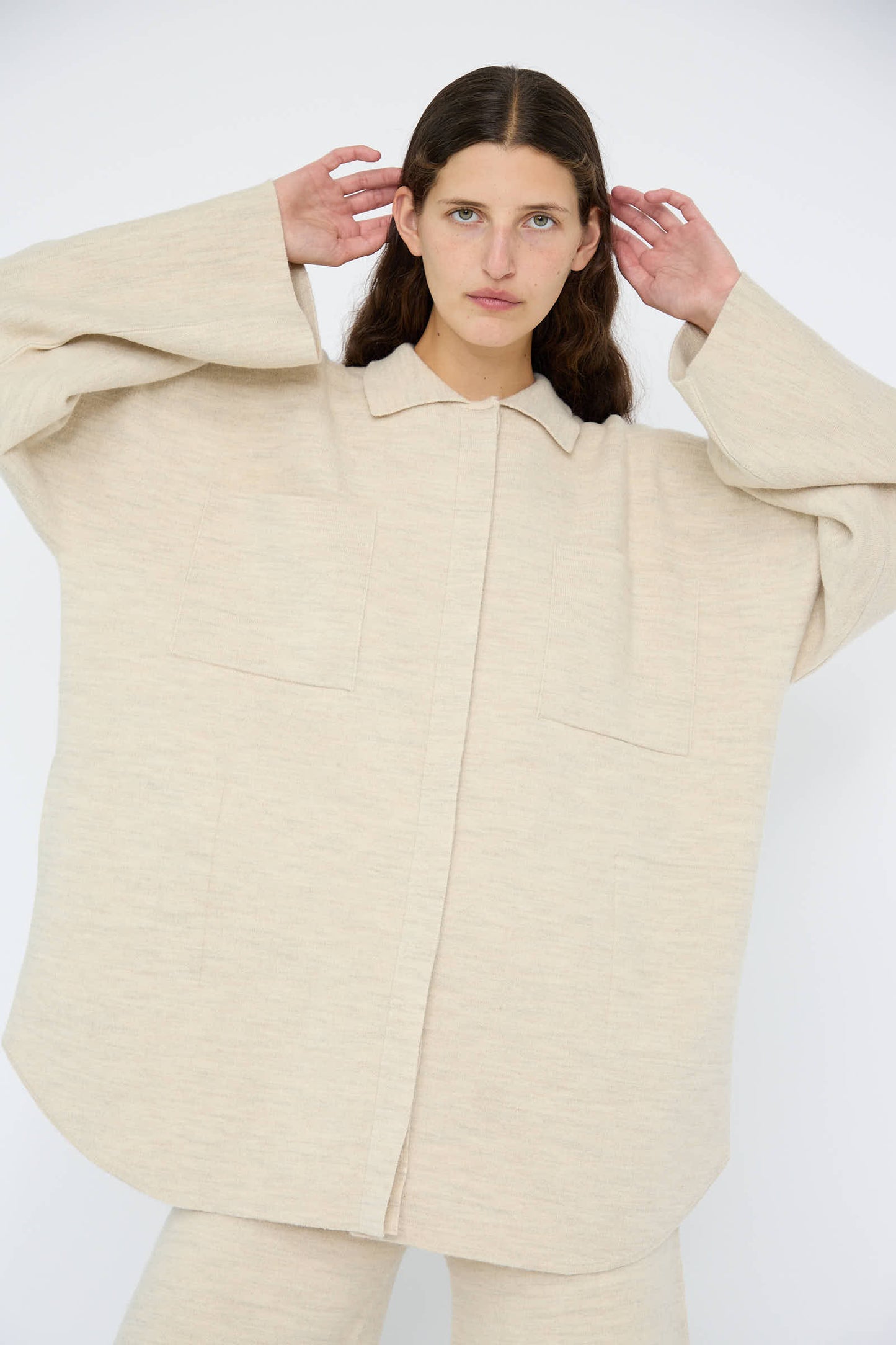 A model dressed in Lauren Manoogian's Double Knit Big Shirt in Ecru, designed with an oversized fit and crafted from a premium wool, alpaca, and cotton blend made in Peru, paired with matching pants against a plain background. Their hands are raised near their head.