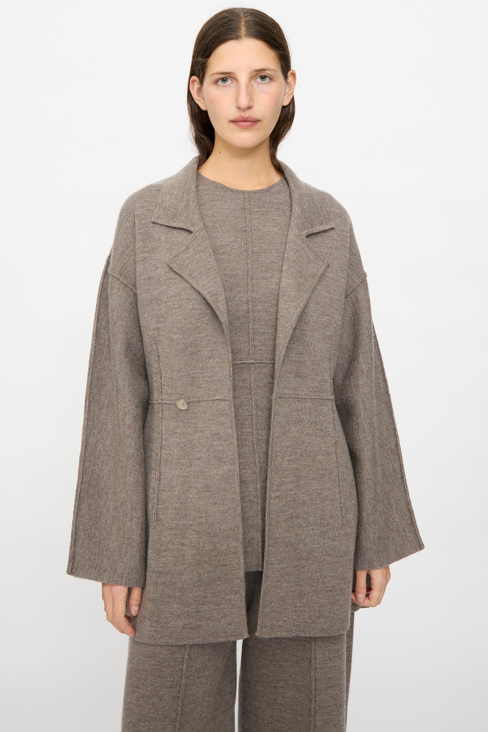 A person stands facing forward in Lauren Manoogian's Double Knit Blazer in Cinder, evoking the sleek silhouette of a women's structured blazer against a plain white background.