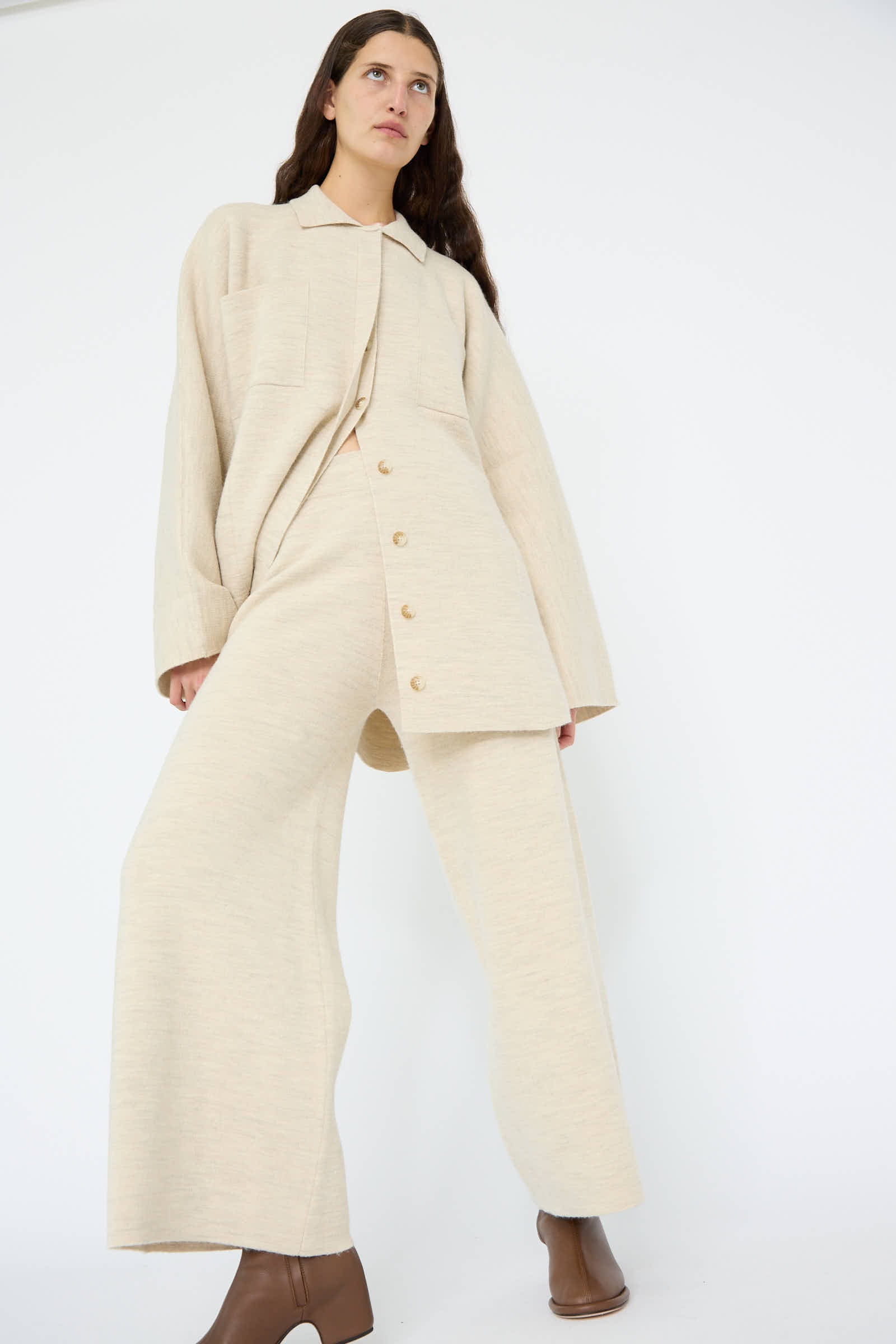 A person stands against a plain white background, wearing an oversized merino wool ensemble with buttons, complemented by the Lauren Manoogian Double Knit Flare Pant in Ecru and brown ankle boots.