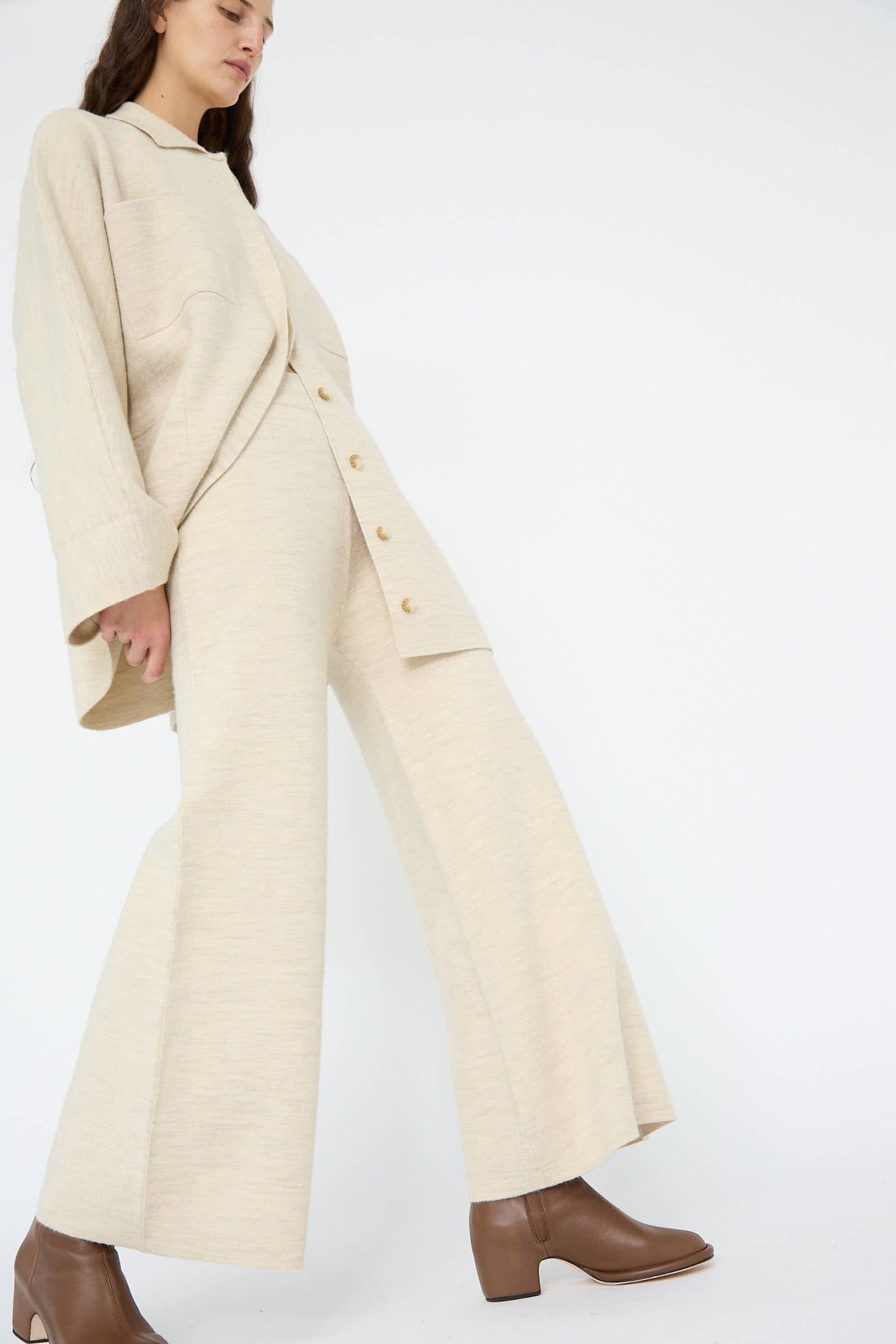 A person dressed in the Double Knit Flare Pant in Ecru by Lauren Manoogian, accompanied by brown ankle boots, is standing against a plain white background.