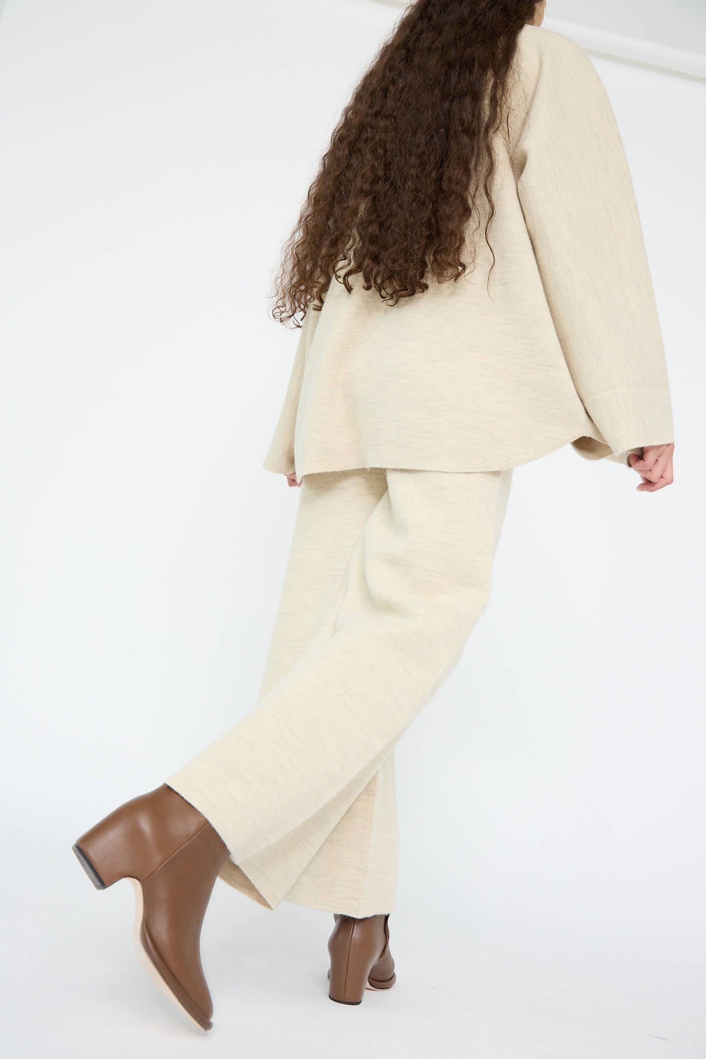 A person in Lauren Manoogian's ecru Double Knit Flare Pant with long hair walks confidently, styled with brown block-heeled ankle boots against a plain white background.