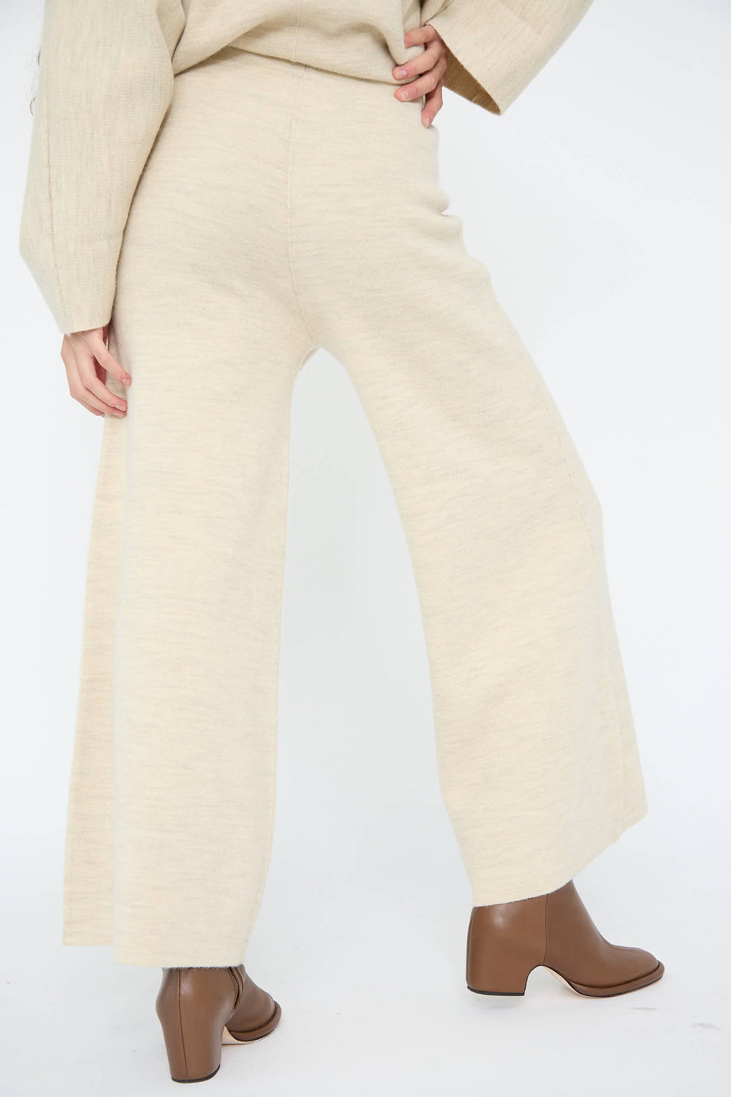 A person is dressed in the Lauren Manoogian Double Knit Flare Pant in Ecru, complemented by a matching top and brown ankle boots, striking a pose with one hand on their hip against a white background.