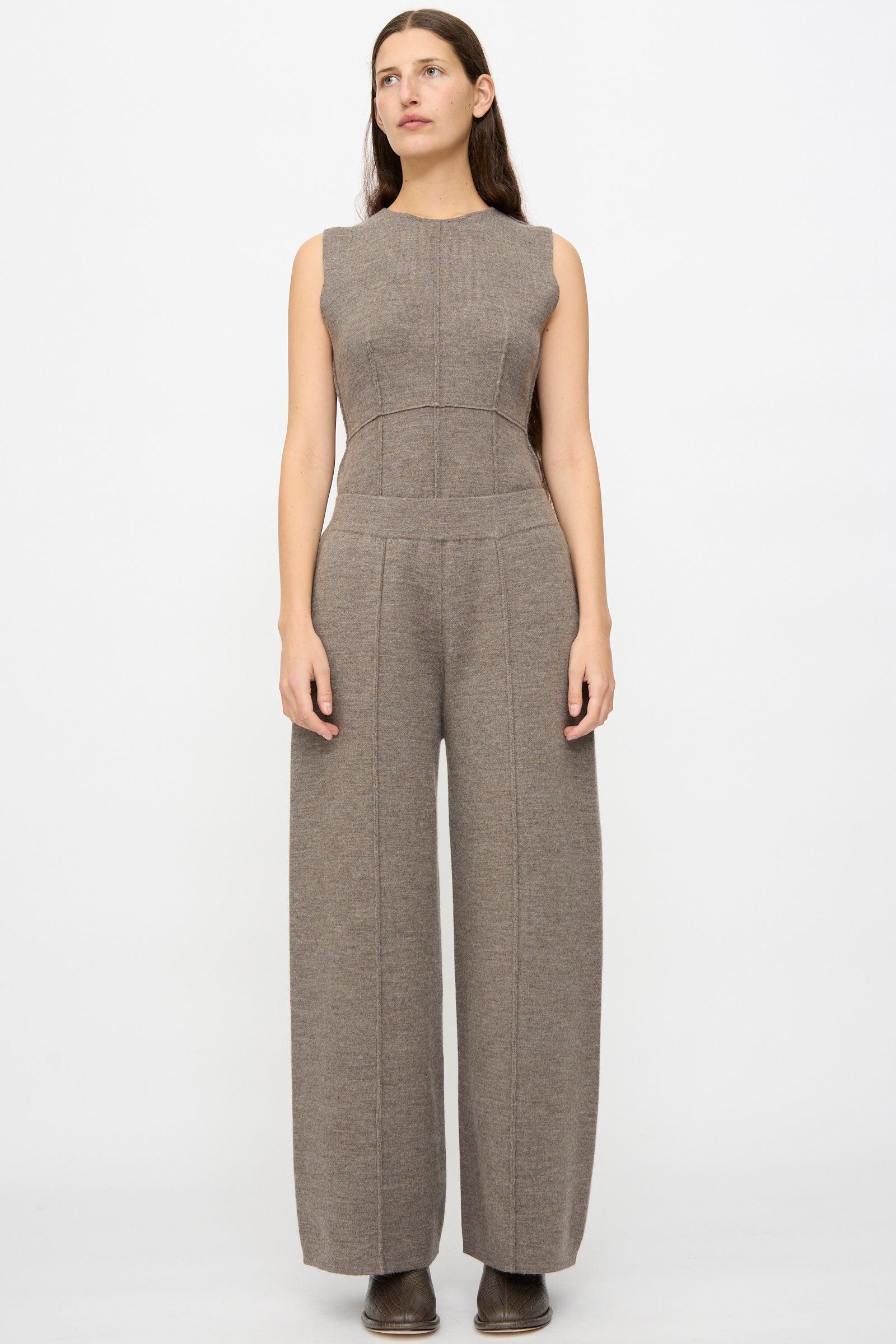 A woman faces forward in Lauren Manoogian’s Double Knit Trouser in Cinder, a sleeveless, wide-leg brown jumpsuit made from soft merino wool, paired with brown shoes, set against a plain white background.