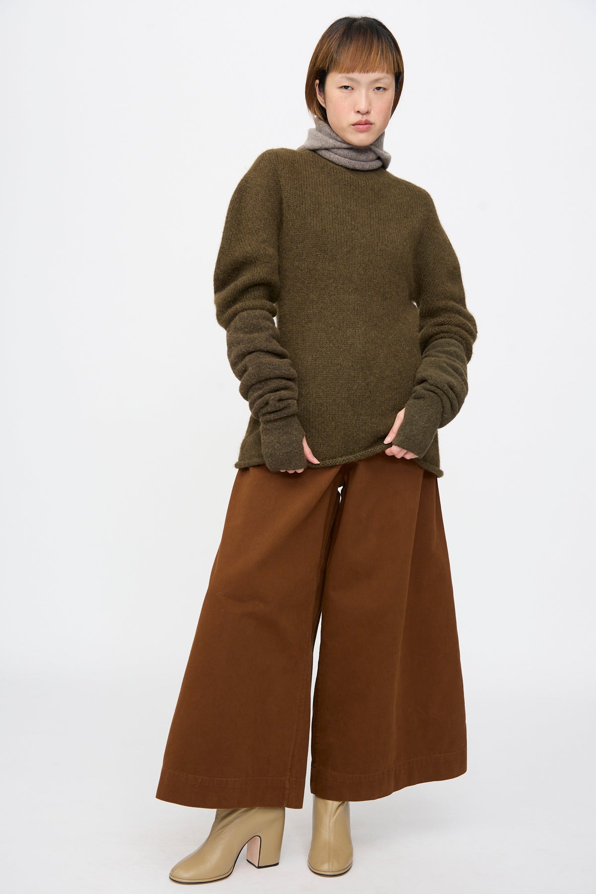 A person wears a baby alpaca blend long-sleeved olive sweater with Lauren Manoogian Felt Fingerless Gloves in Olive, wide-leg brown trousers, and beige boots.