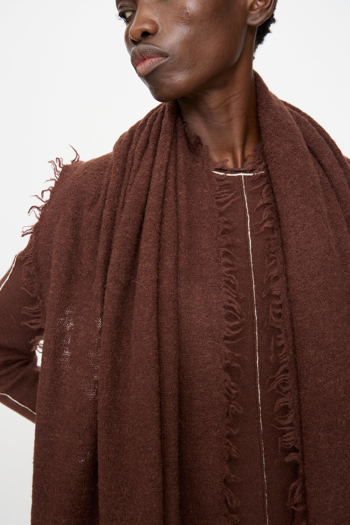 A person wears the Lauren Manoogian Felt Fringe Scarf in Merlot—an oversized, textured brown merino wool scarf with frayed edges—styled with a matching top, shown from the shoulders up against a plain background.
