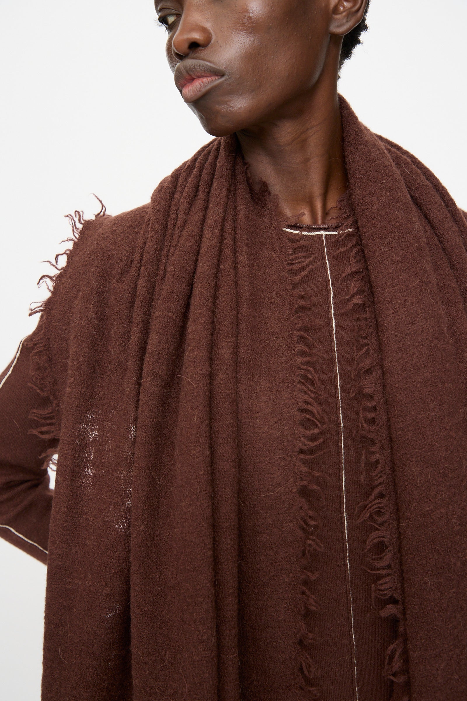 A person wears the Lauren Manoogian Felt Fringe Scarf in Merlot—an oversized, textured brown merino wool scarf with frayed edges—styled with a matching top, shown from the shoulders up against a plain background.
