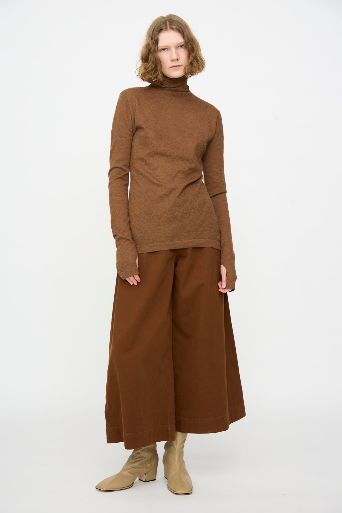 A person stands against a plain white background, wearing the Fine Funnel in Rust turtleneck by Lauren Manoogian, paired with wide brown culottes and beige boots, with shoulder-length wavy hair.