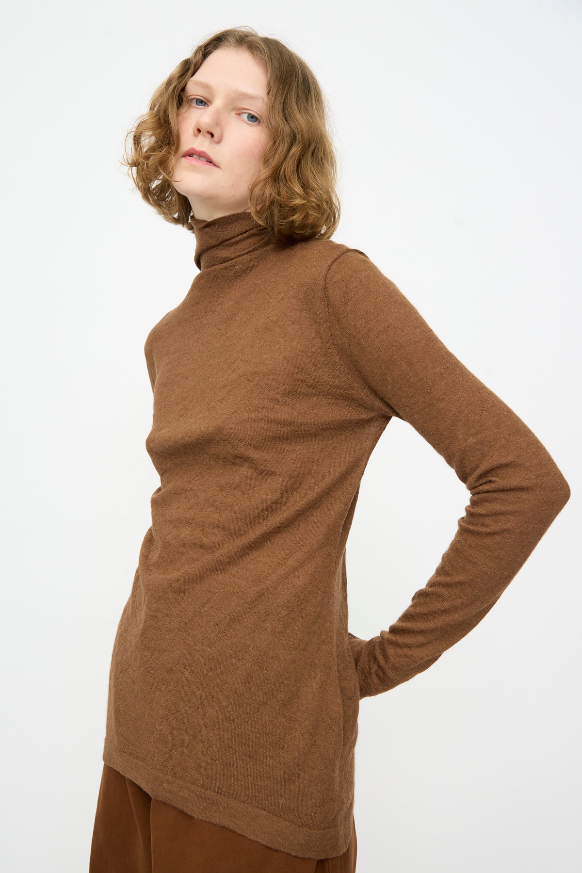 A person with short curly hair wears the Lauren Manoogian Fine Funnel sweater in Rust—crafted from baby alpaca and made in Peru—paired with matching brown pants, standing against a plain white background.