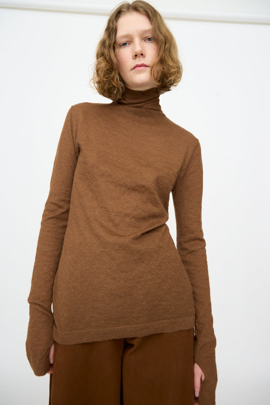 A person with shoulder-length curly hair wears the Lauren Manoogian Fine Funnel in Rust and brown pants, standing against a plain white background.