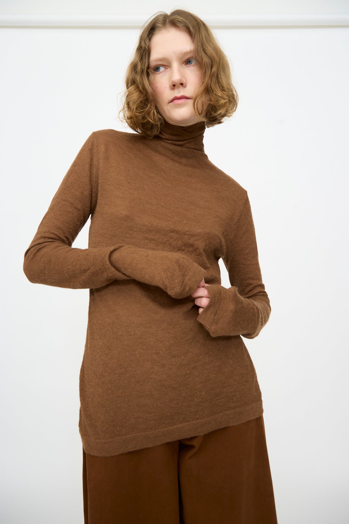 A person with short curly hair wearing the Lauren Manoogian Fine Funnel in Rust and brown pants stands against a plain white background.
