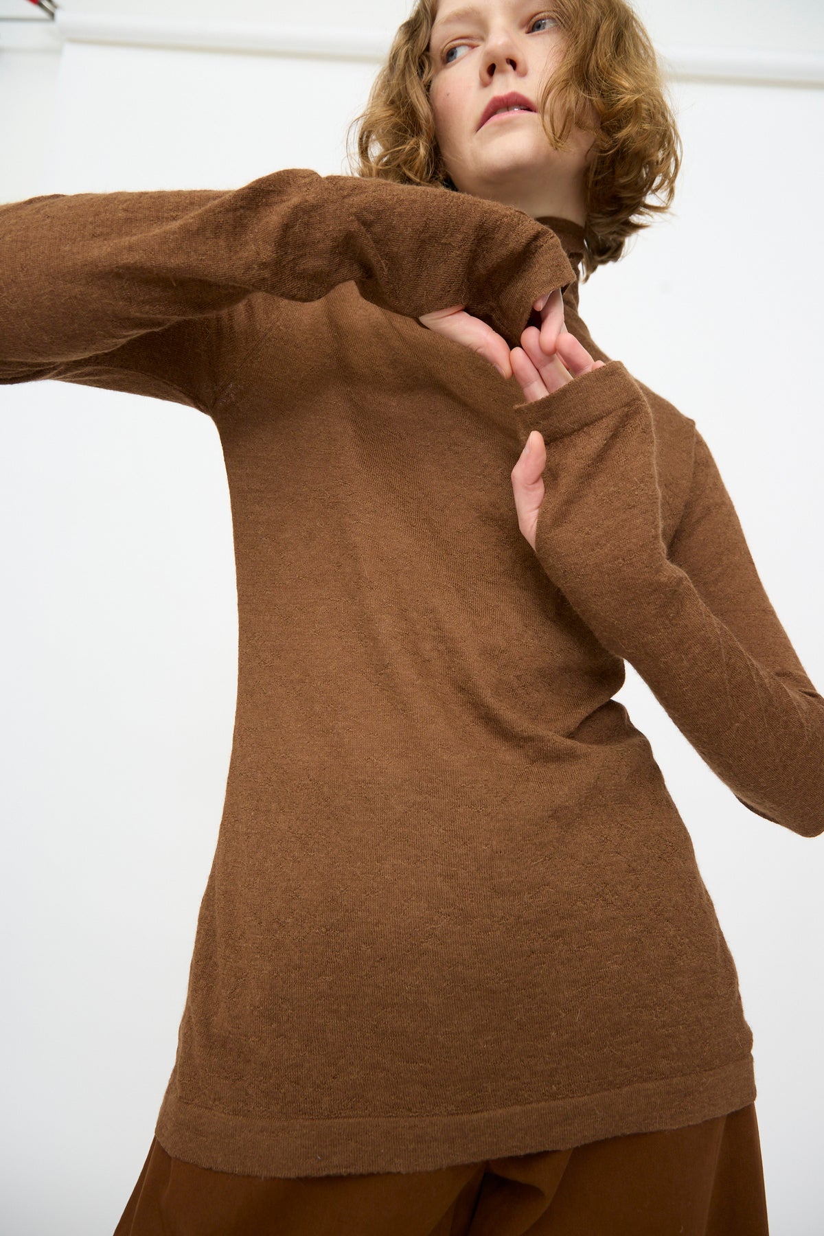 A person wears the Lauren Manoogian Fine Funnel in Rust, paired with matching brown pants, standing against a plain white background with arms bent and one hand near their face.