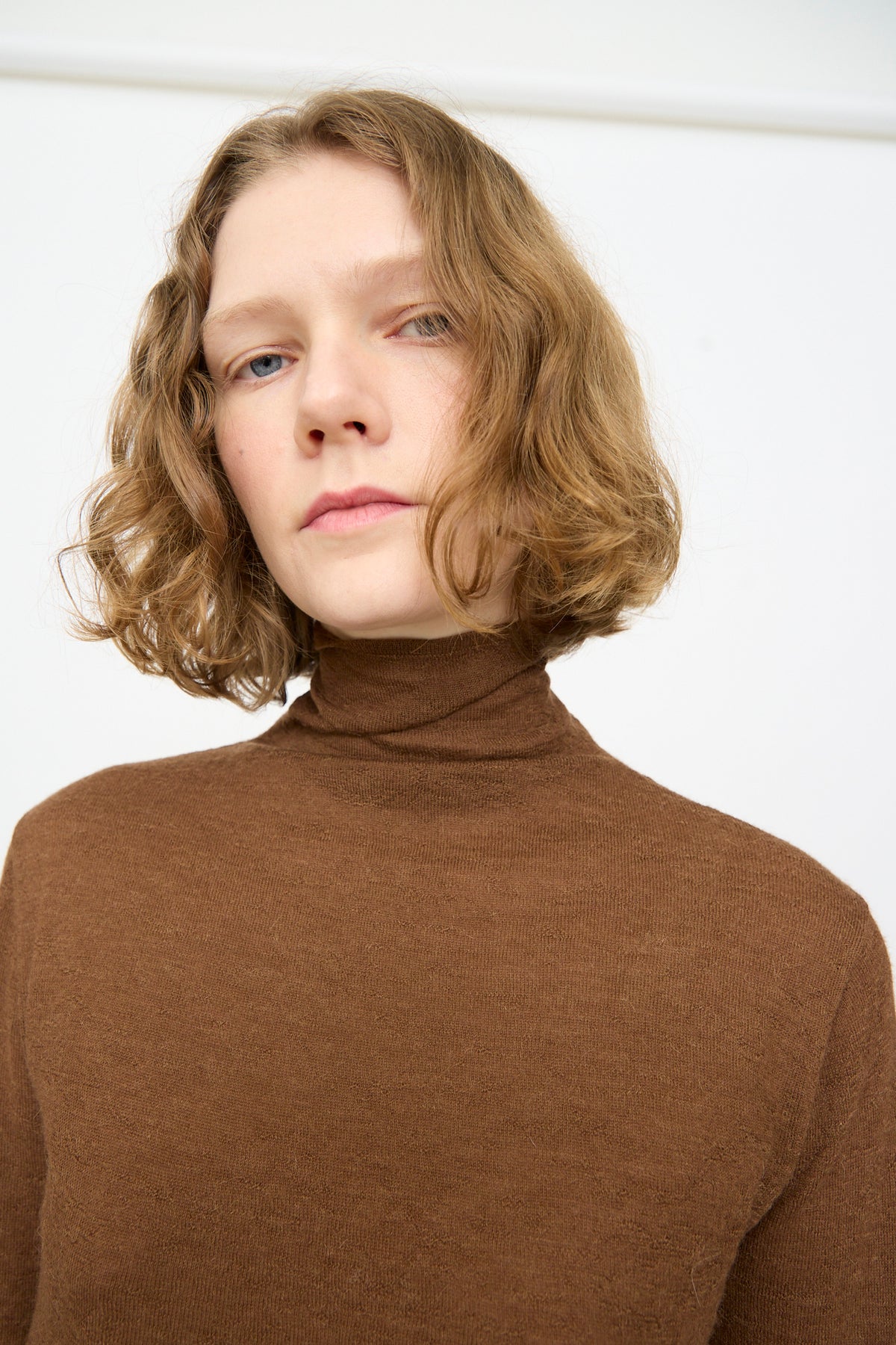 A person with short, wavy, light brown hair wears the Lauren Manoogian Fine Funnel in Rust and looks at the camera against a plain white background.