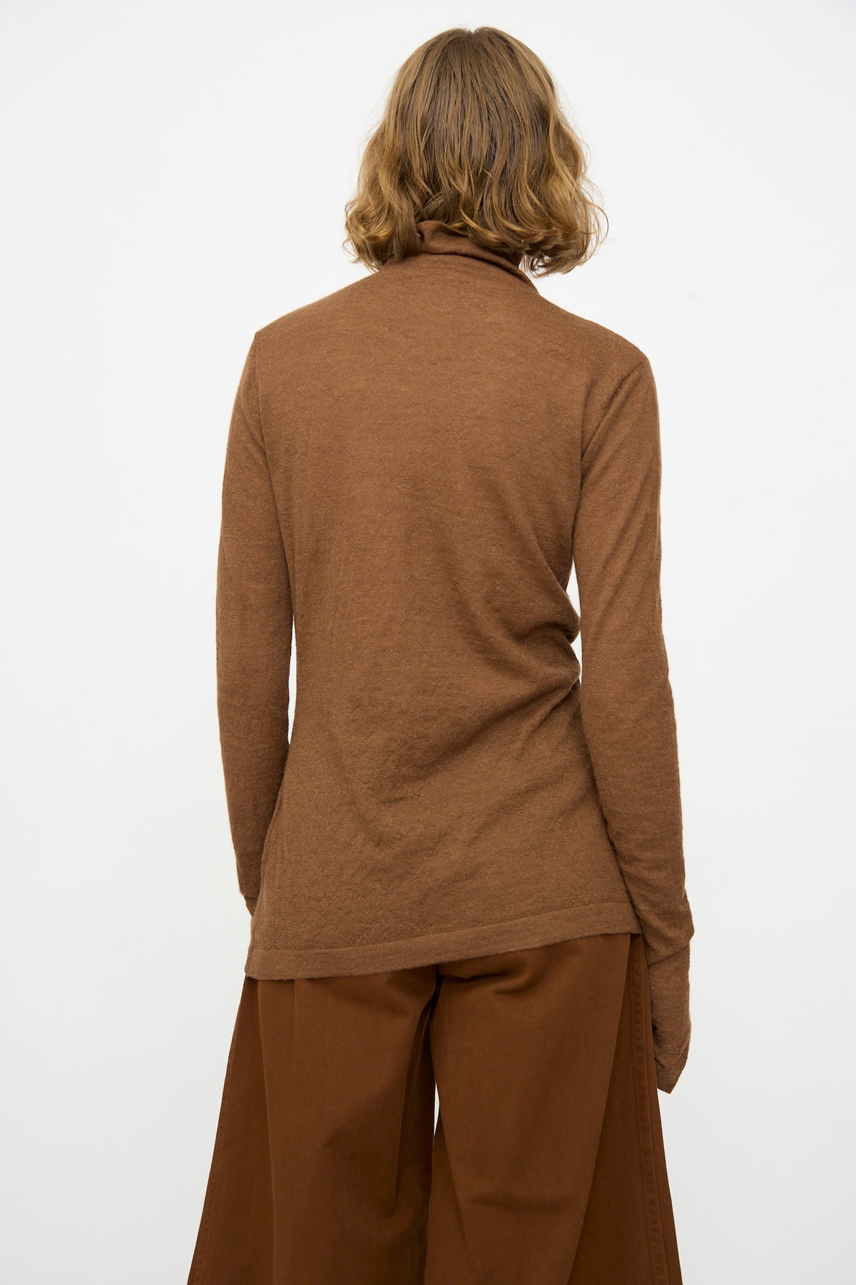 A person with shoulder-length hair faces away from the camera, wearing the Lauren Manoogian Fine Funnel in Rust sweater and wide brown pants against a plain white background.