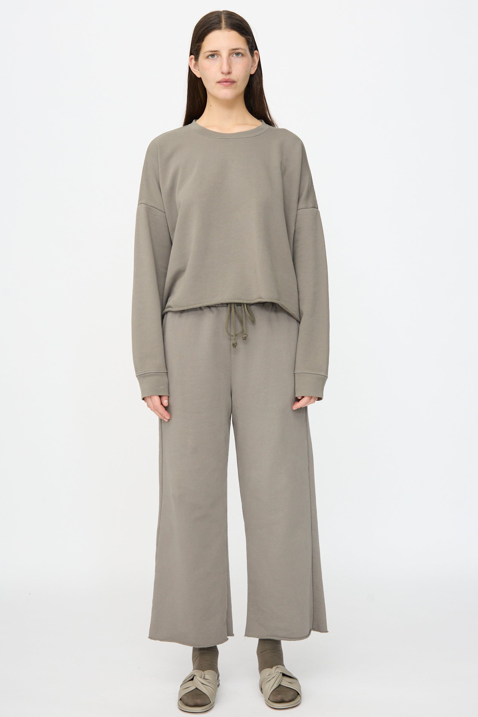 A person faces forward in Lauren Manoogian Fleece Pants in Fatigue with an elastic waist and a matching loose-fit sweatshirt, plus beige sandals and socks. The background is plain white.
