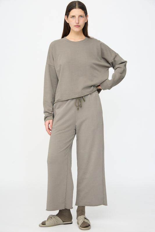 A person stands facing forward in Lauren Manoogian Fleece Pants in Fatigue, a loose-fitting taupe sweatshirt, taupe socks, and slide sandals against a plain white background.