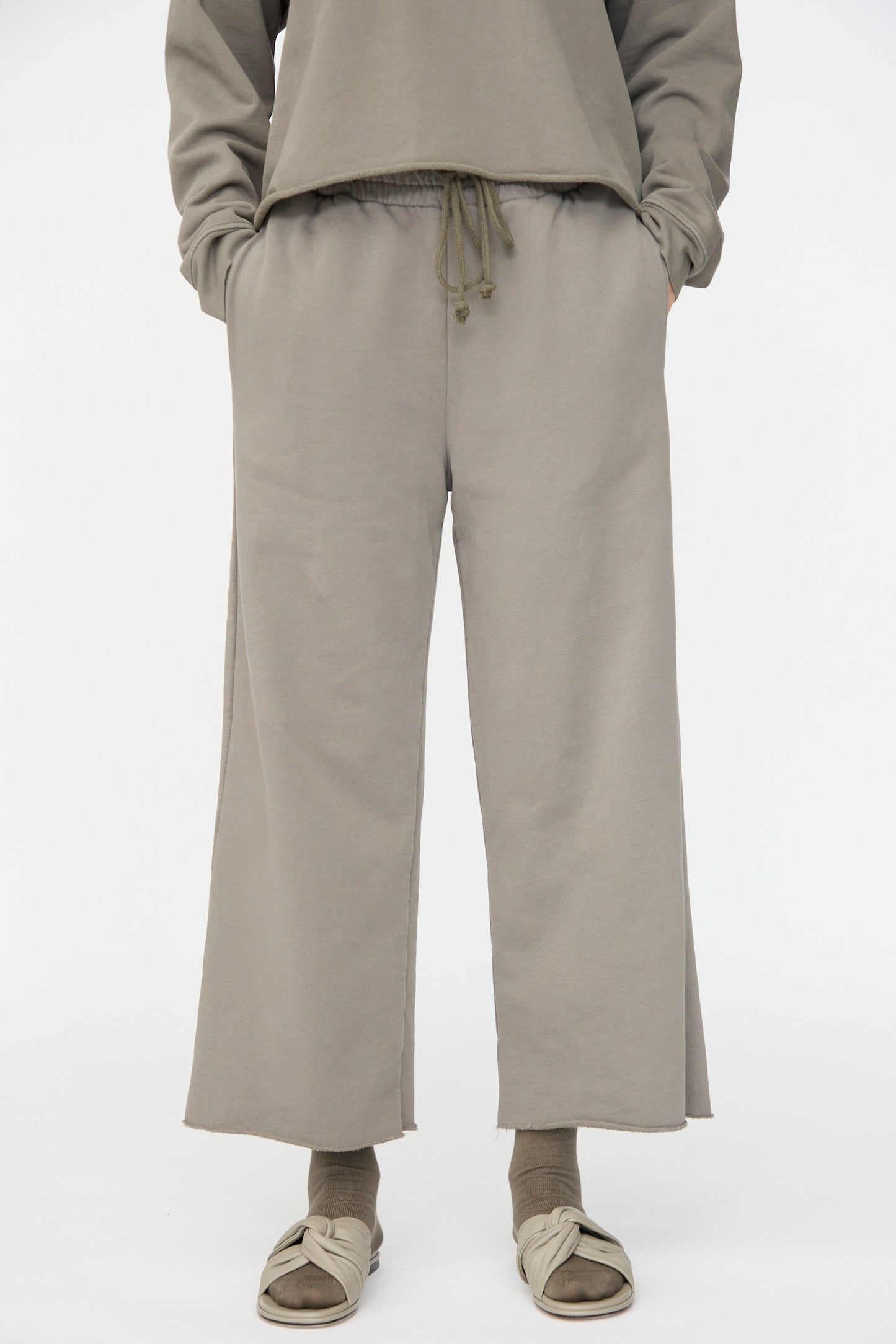 Person wearing Lauren Manoogian Fleece Pants in Fatigue, featuring a relaxed fit and elastic waist, paired with matching socks and beige slide sandals; hands in pockets.