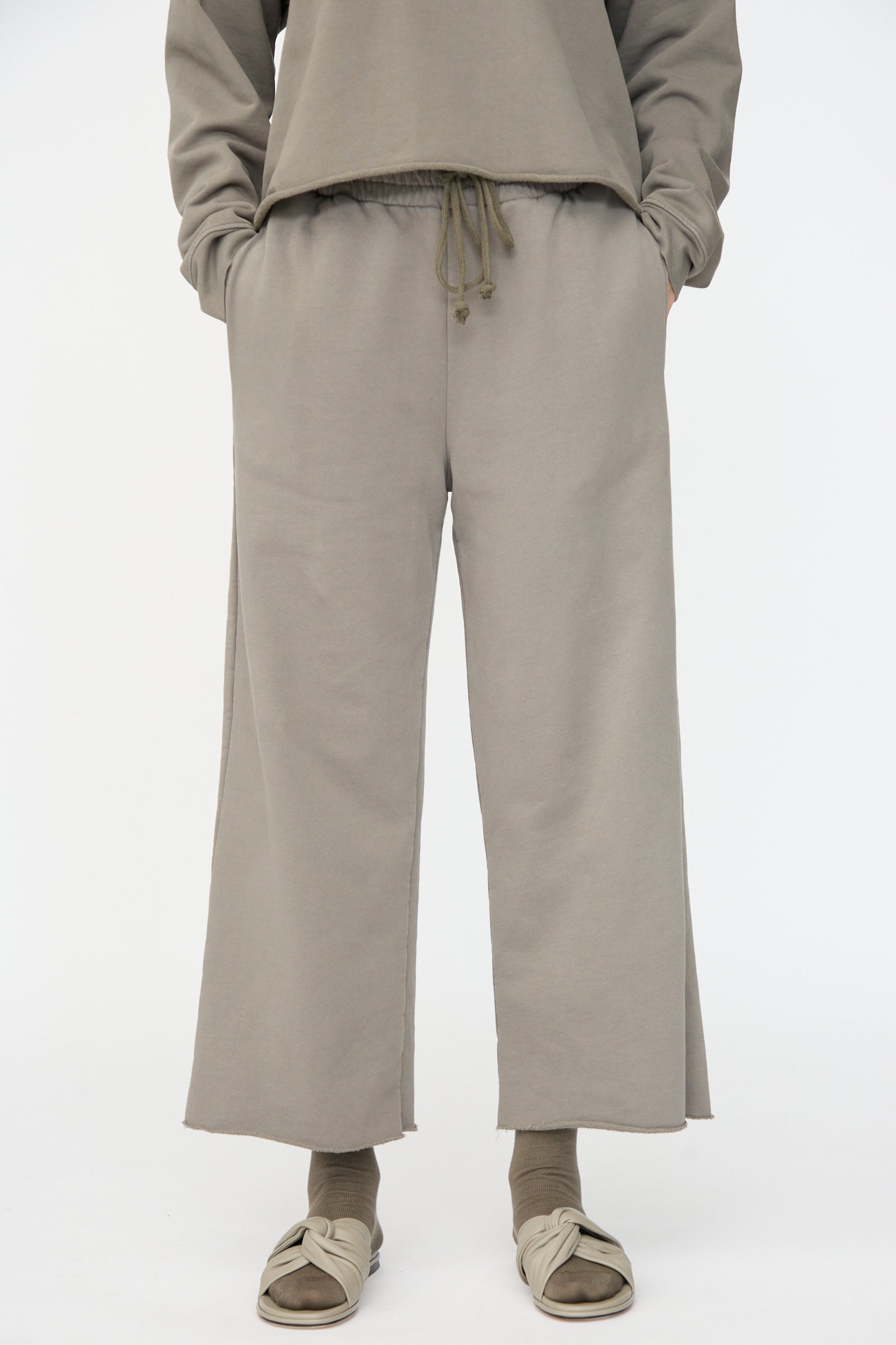 Person wearing Lauren Manoogian Fleece Pants in Fatigue, featuring a relaxed fit and elastic waist, paired with matching socks and beige slide sandals; hands in pockets.