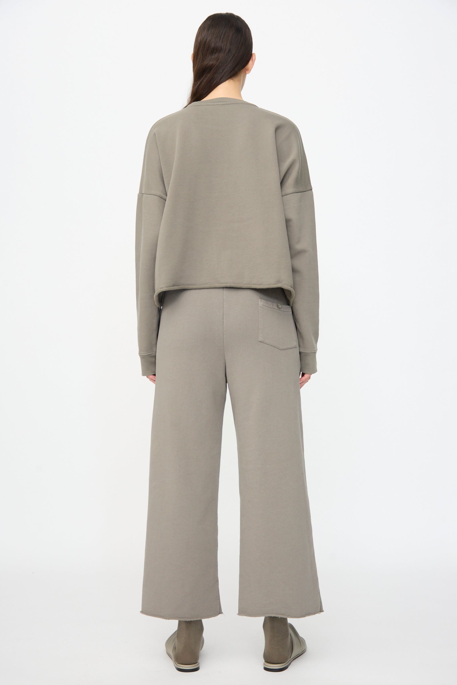 A person stands facing away in Lauren Manoogian Fleece Pants in Fatigue—relaxed fit with elastic waist—paired with a loose taupe sweatshirt and matching shoes, all set against a plain white background.