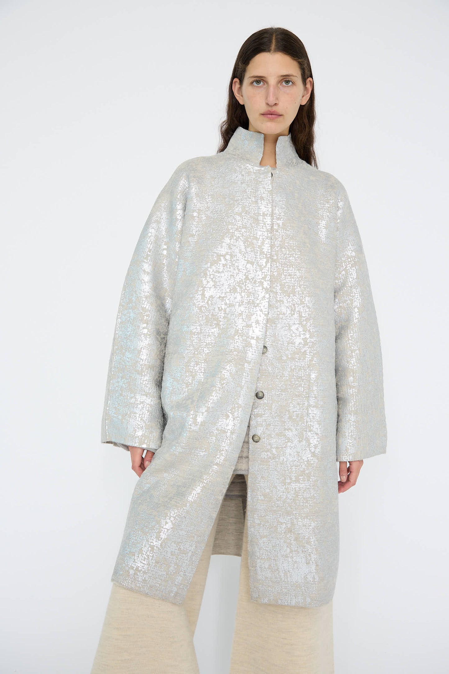 A person wearing the Foil Car Coat in Silver Carrara by Lauren Manoogian, paired with wide beige pants, stands against a plain background.