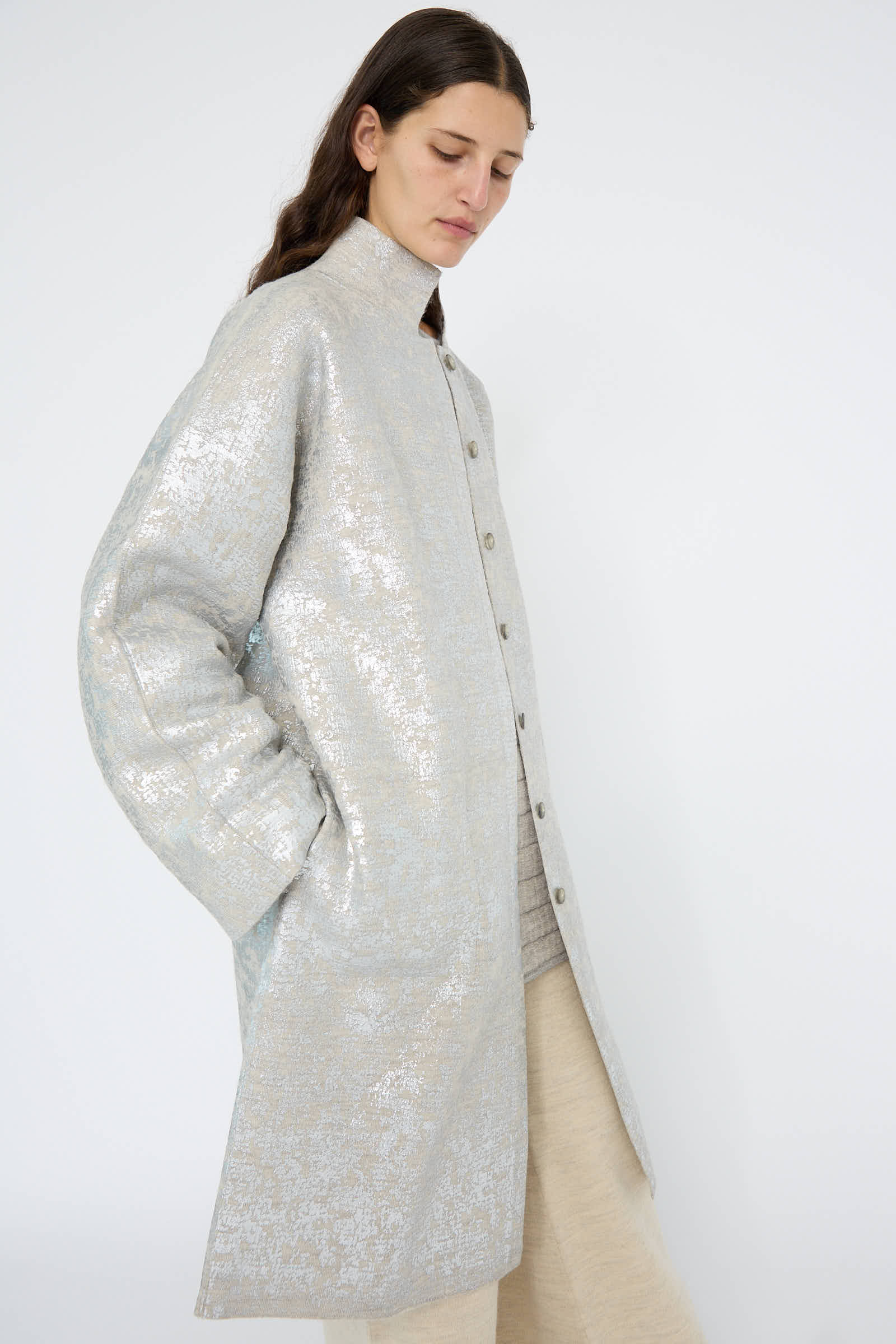 A person wears the Lauren Manoogian Foil Car Coat in Silver Carrara over a light outfit, gazing downward against a plain background.