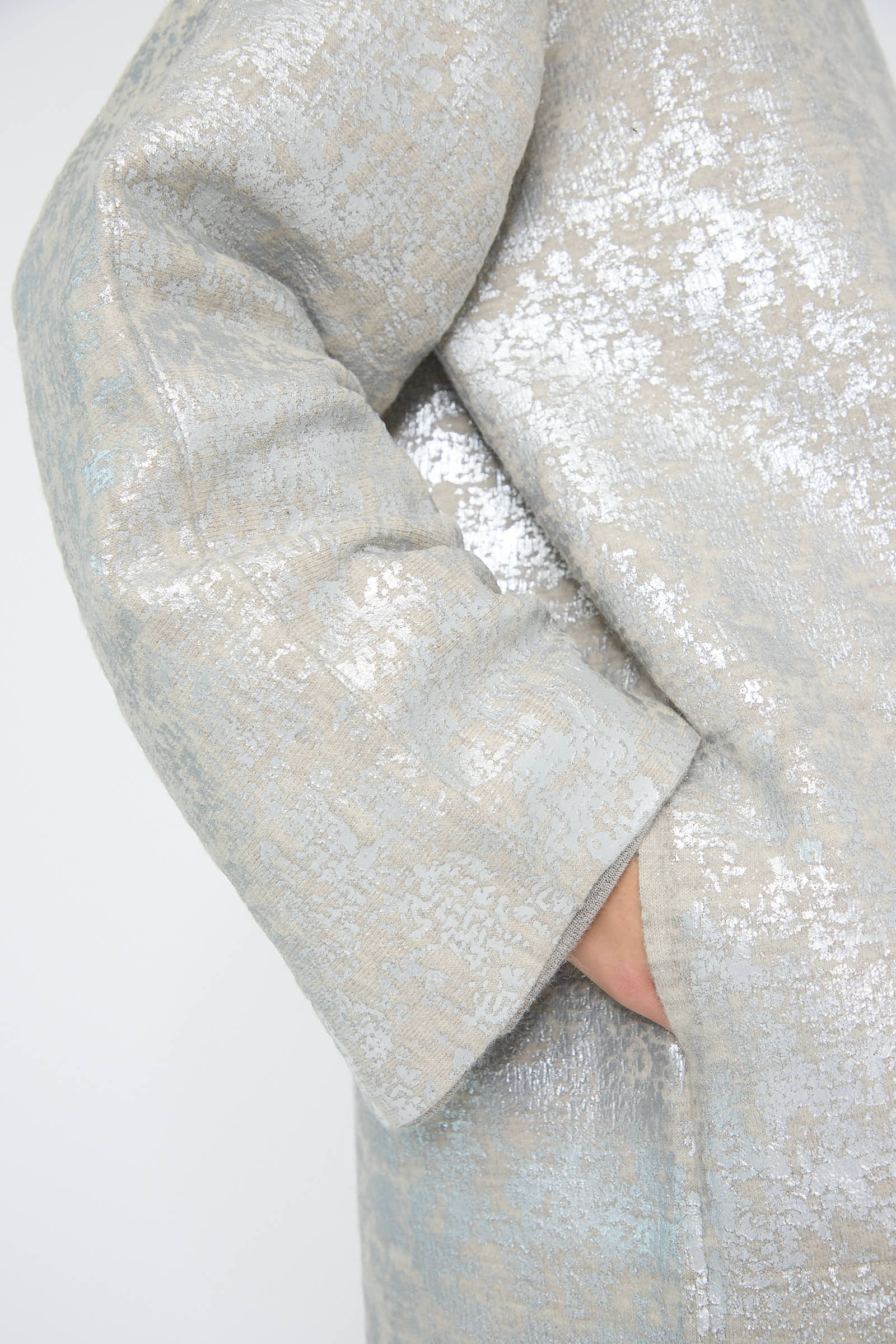 A close-up of a person’s arm in the textured, shimmering "Foil Car Coat in Silver Carrara" by Lauren Manoogian, featuring a hand partially tucked into the side pocket.