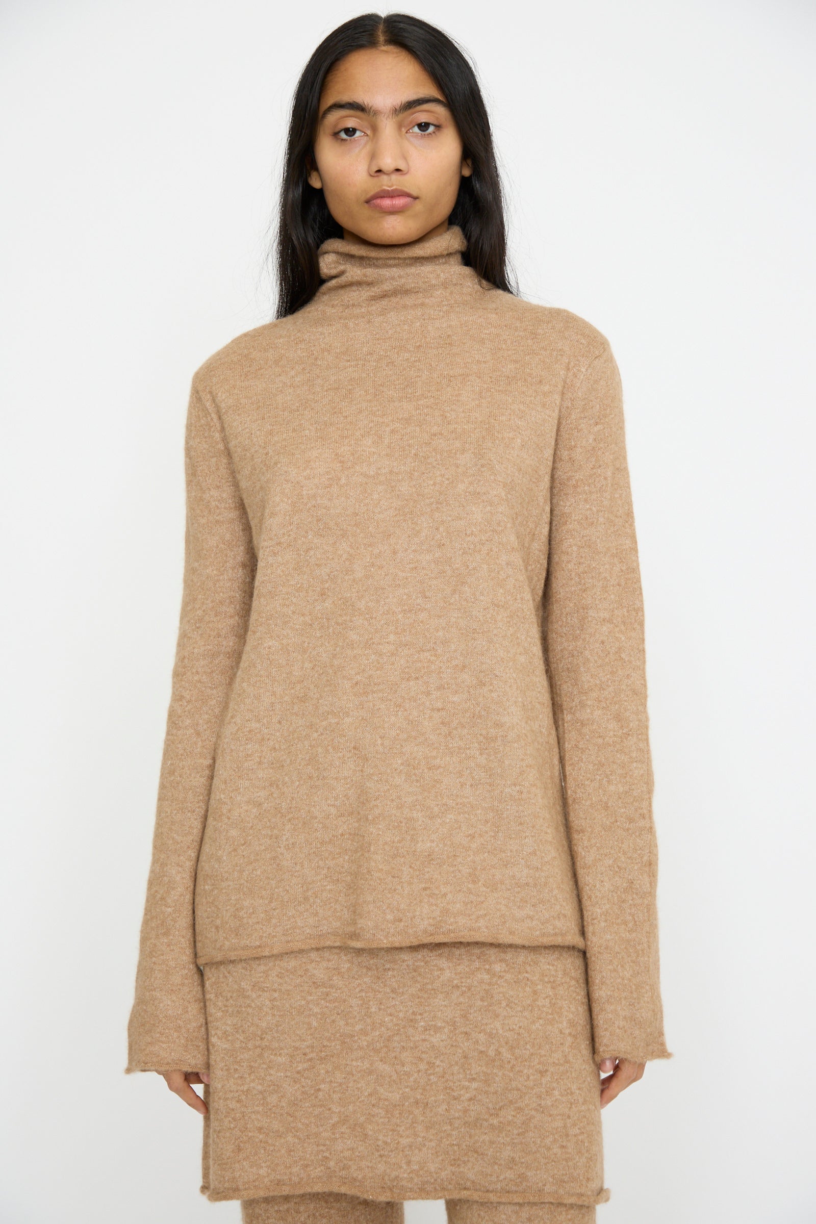 A person wears a camel Funnel Turtleneck by Lauren Manoogian and a matching skirt, standing against a plain backdrop.